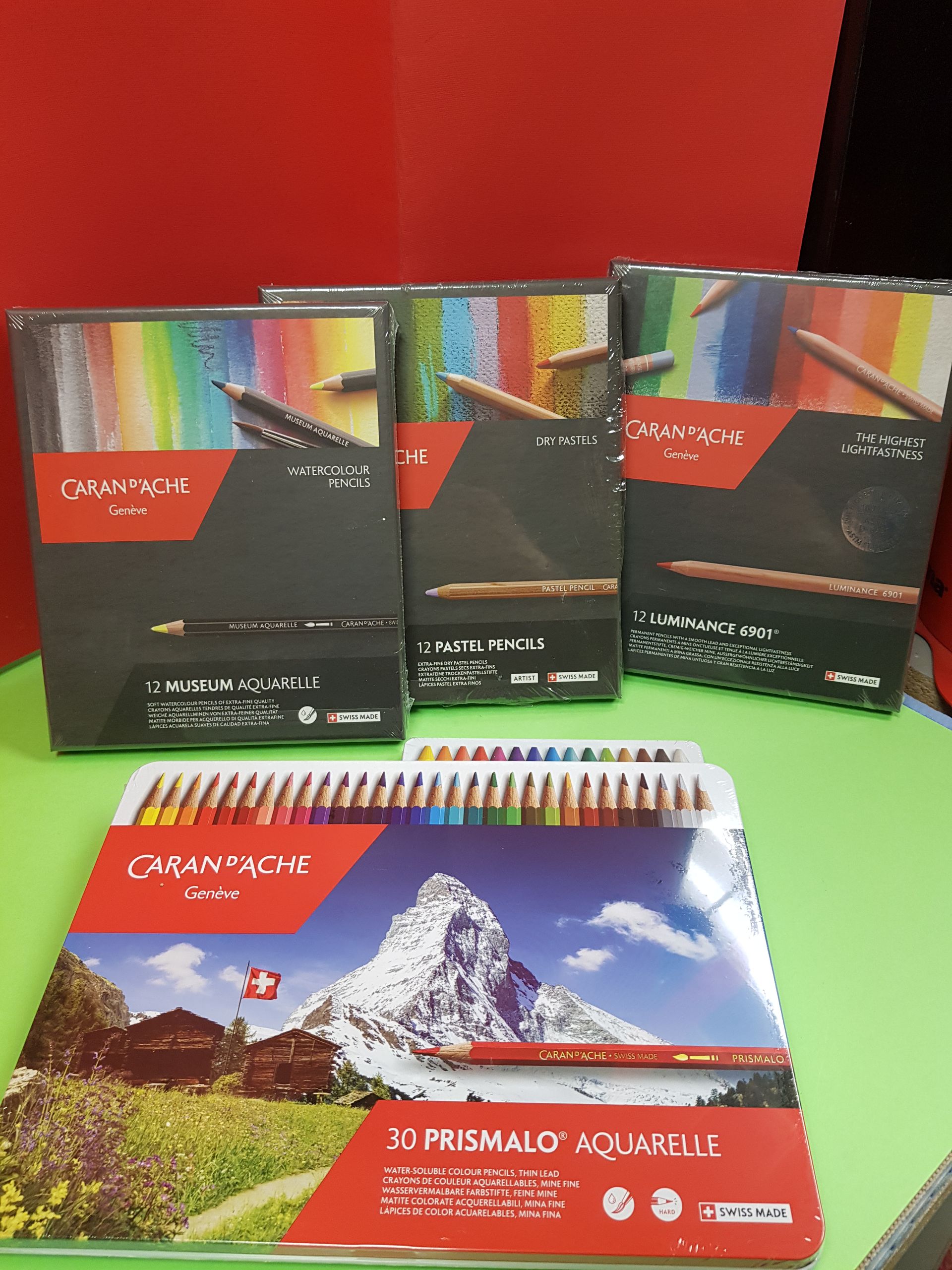 Scatole di matite colorate su una superficie verde, con un paesaggio montano svizzero sulla parte anteriore di una.