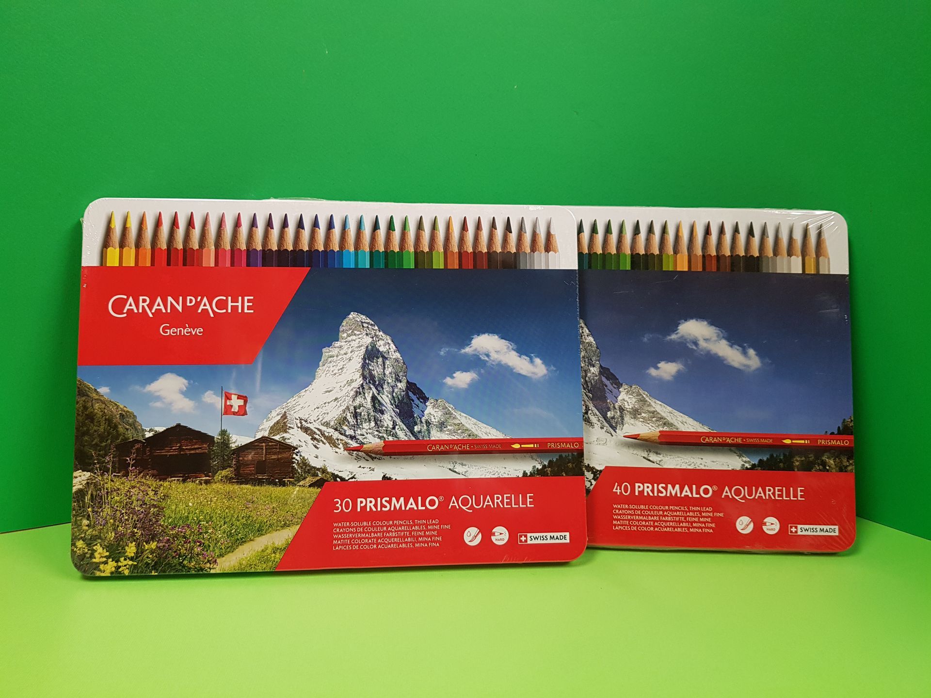 Due set di matite acquerellabili Caran d'Ache verdi, uno con 30 matite e l'altro con 12, raffiguranti un paesaggio montano.