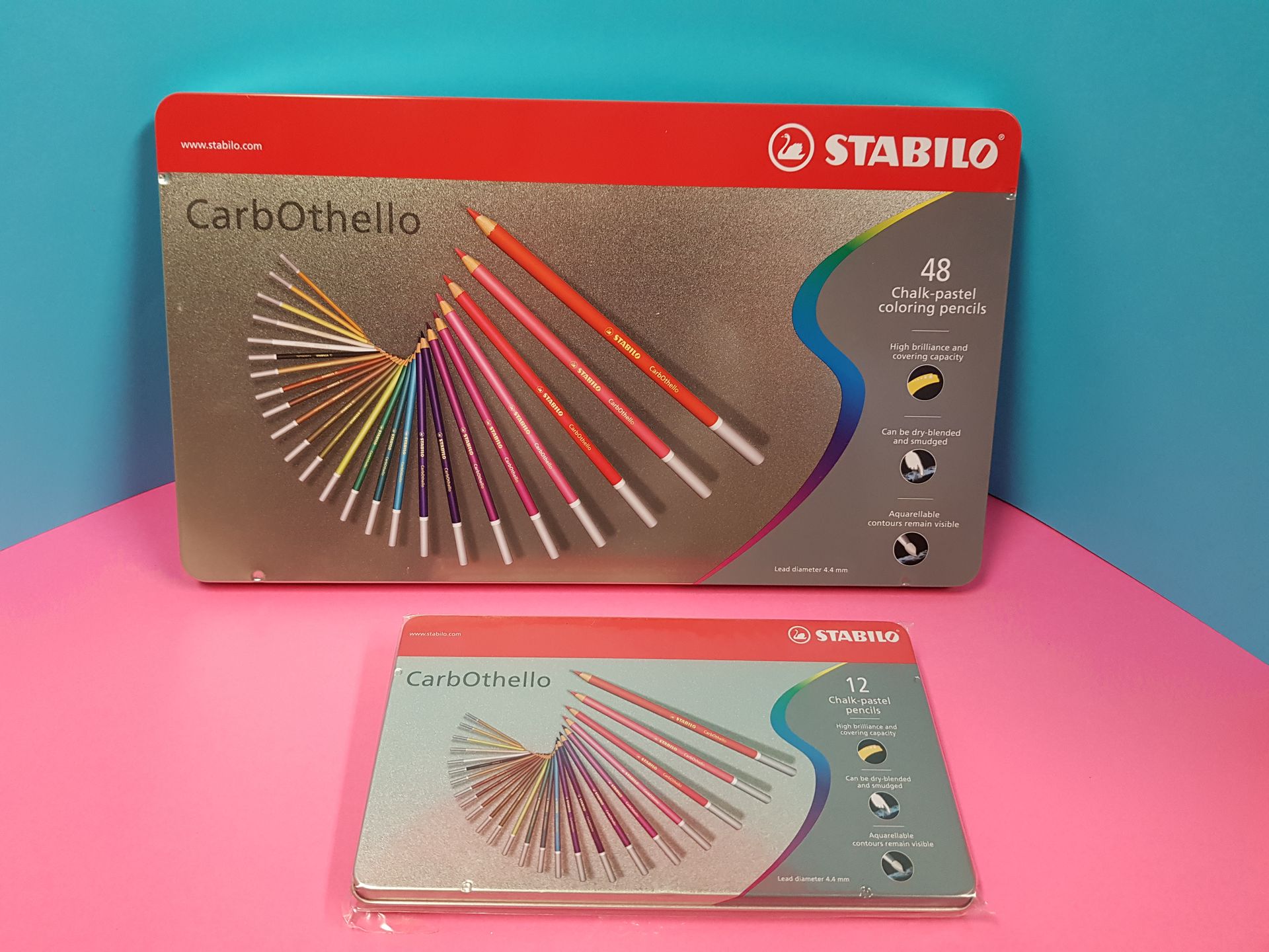 Due set di matite pastello Stabilo CarbOthello, uno grande e uno piccolo, su una superficie rosa con sfondo blu.