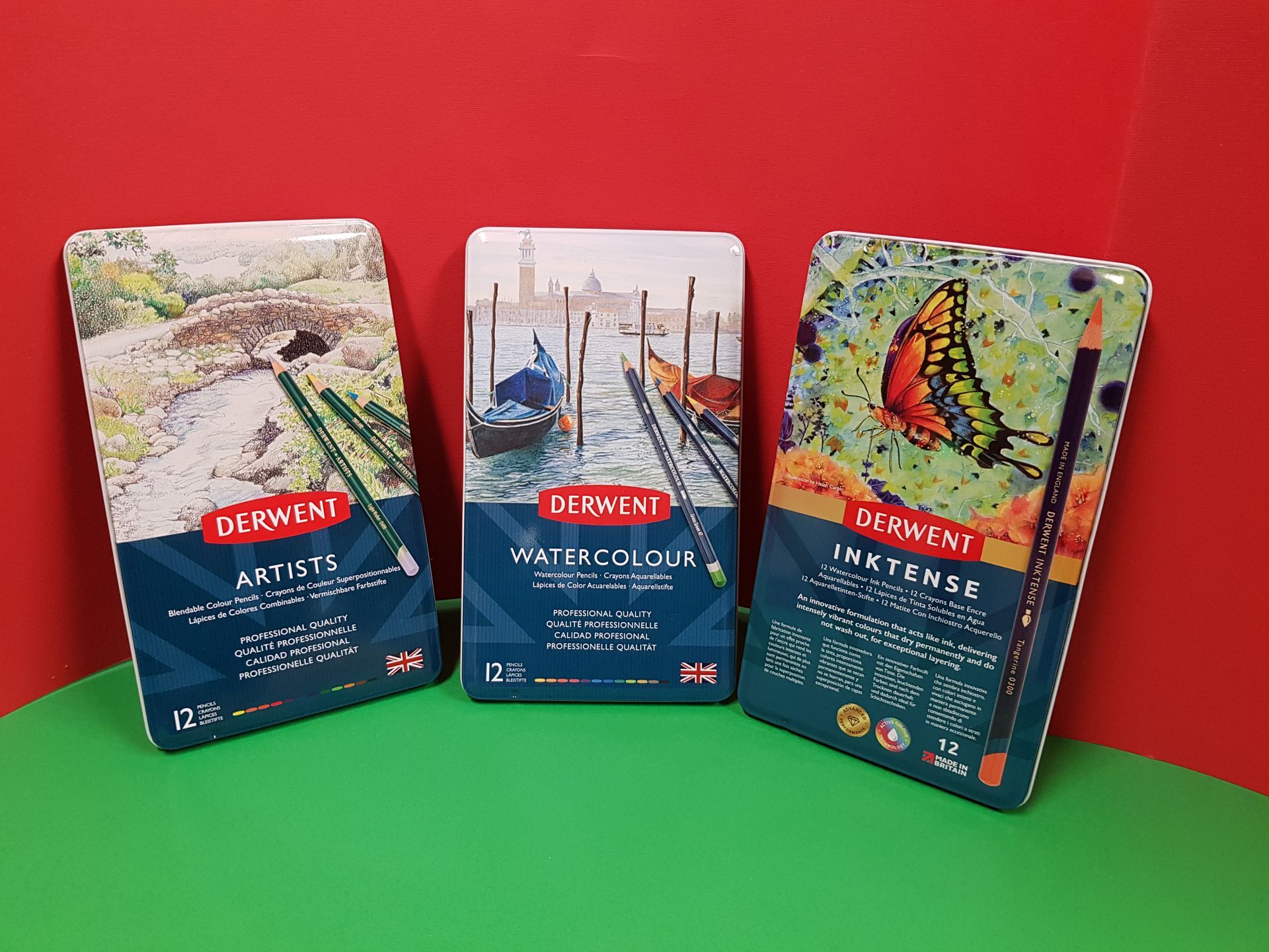 Tre scatole di matite Derwent: Drawing, Watercolour e Inktense, su superficie verde su sfondo rosso.