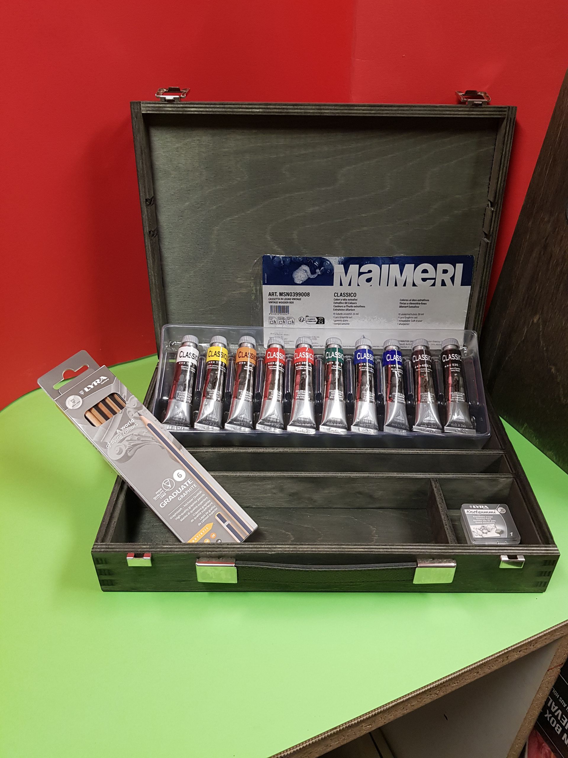 Valigetta in legno aperta da artista contenente tubetti di vernice, matite e un orologio su una superficie verde.