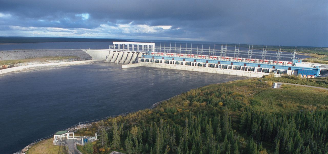 Vue aérienne d'un grand barrage hydroélectrique dans un paysage naturel avec une eau bleu foncé et un ciel nuageux.