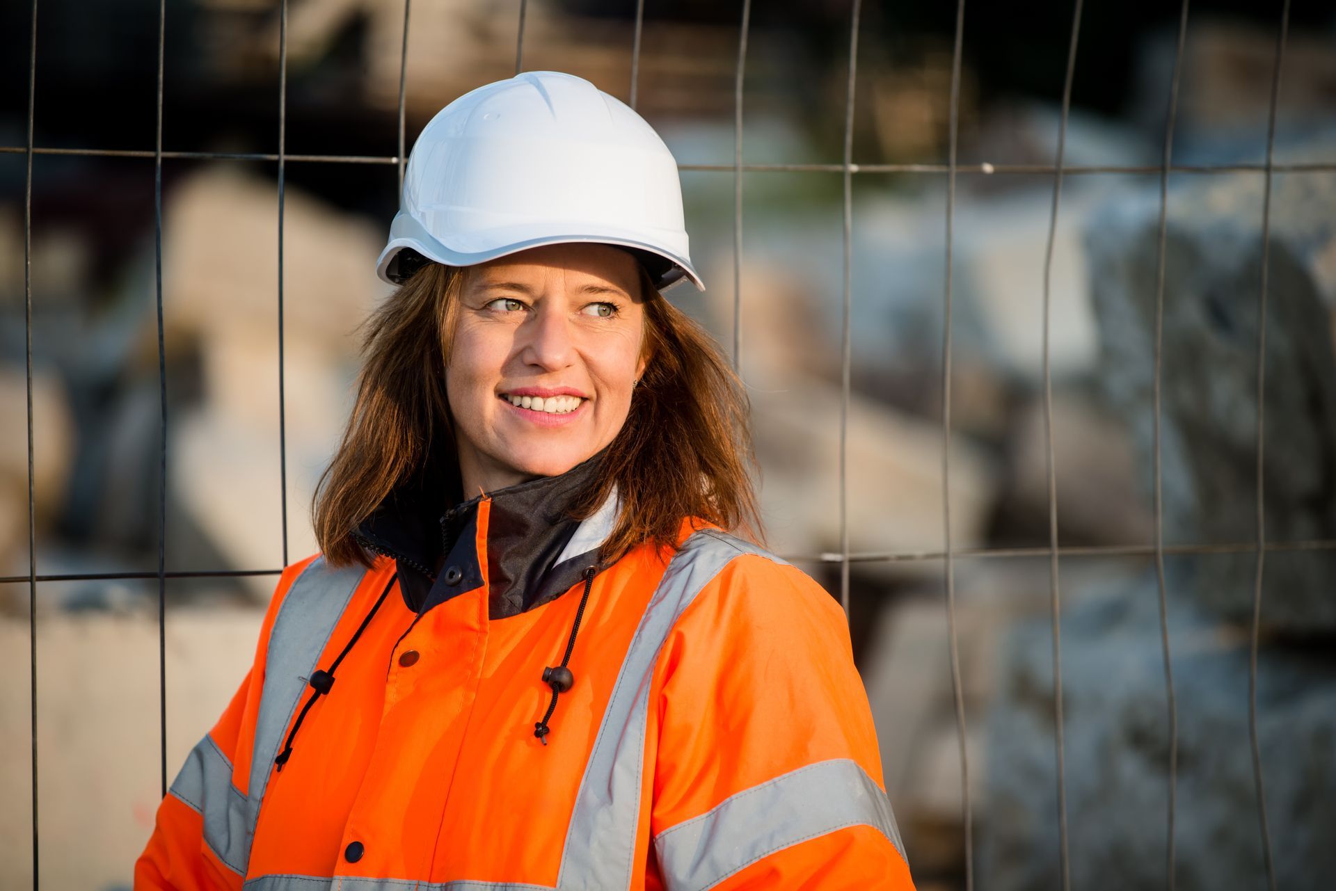 Une femme portant un casque blanc et un gilet de sécurité orange sourit près d'une barrière de chantier.