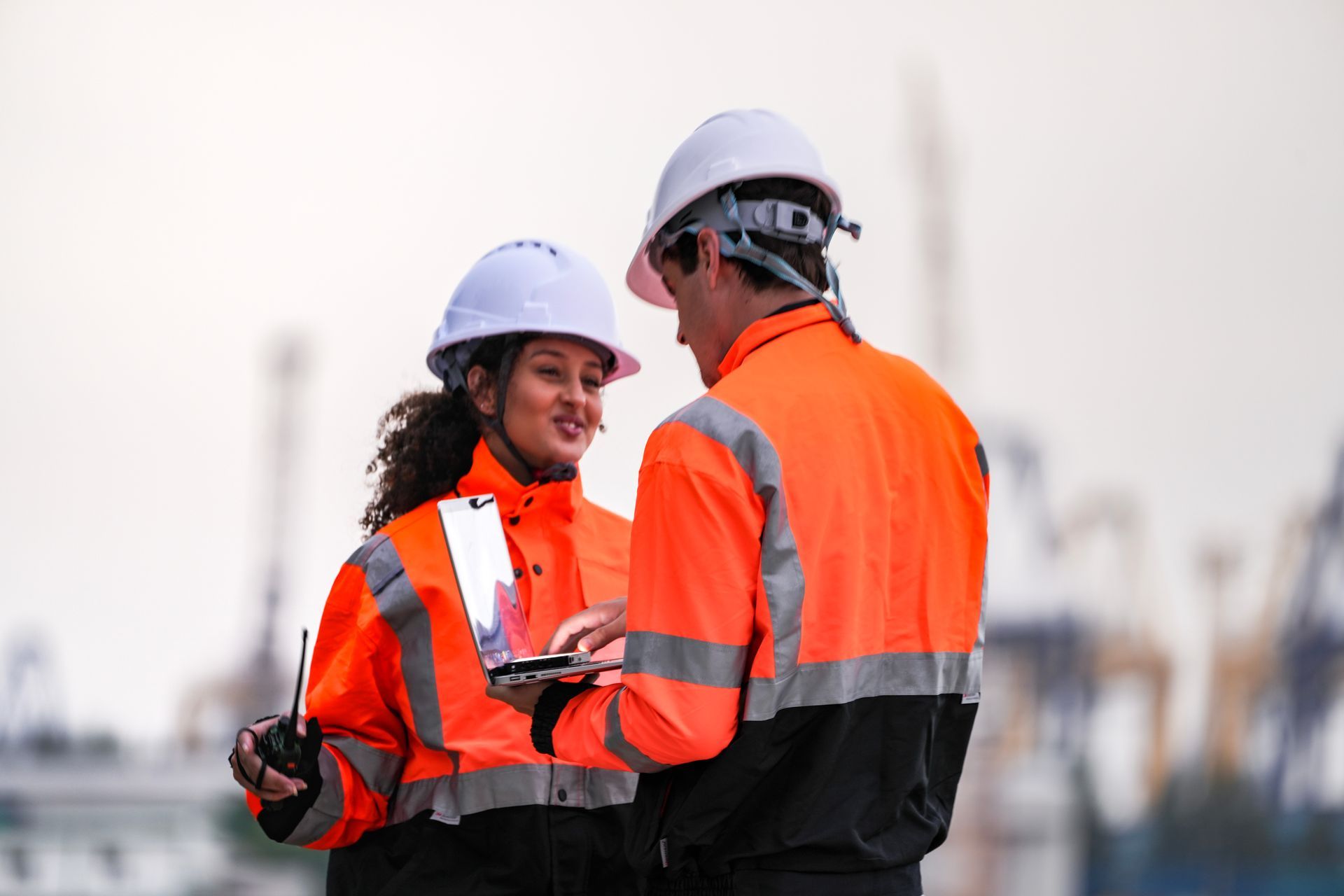 Deux personnes portant des gilets de sécurité et des casques de chantier regardent un ordinateur portable et une radio, à l'extérieur.