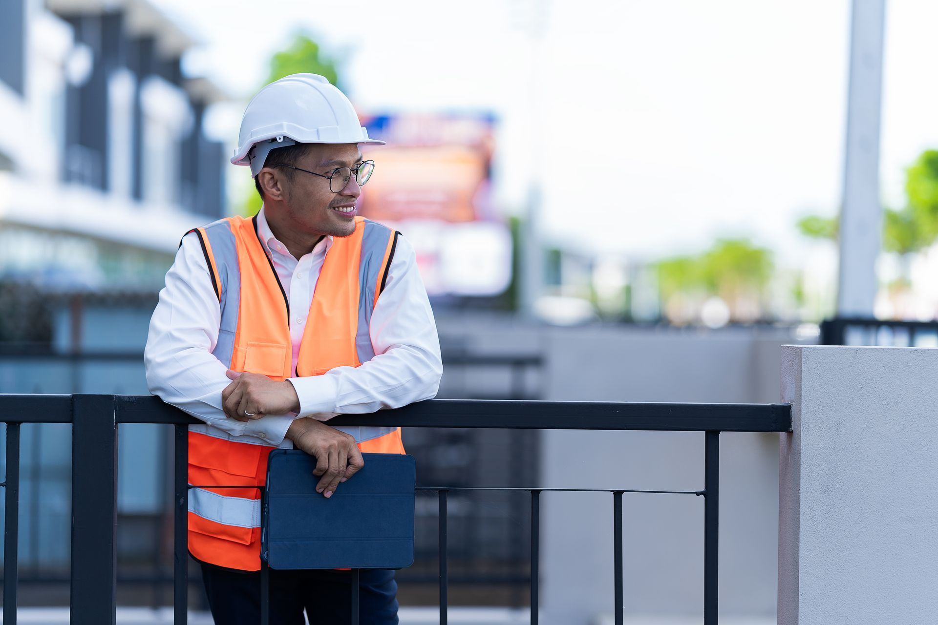 Un ouvrier du bâtiment, portant un casque, des lunettes et un gilet de sécurité, appuyé contre une clôture, tient une tablette.