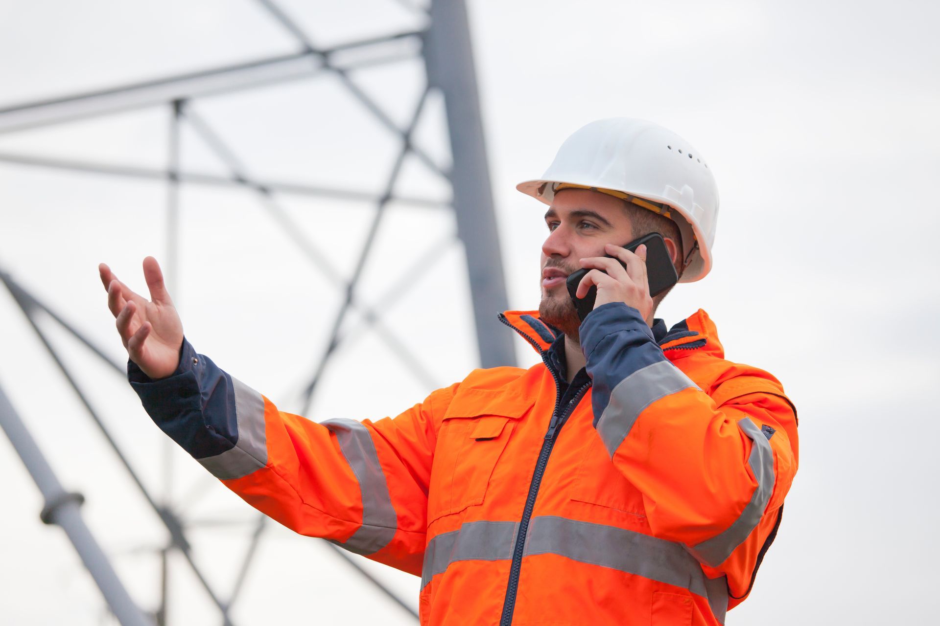 Un homme portant un gilet de sécurité orange et un casque de chantier parle au téléphone près de lignes électriques, en faisant des gestes.