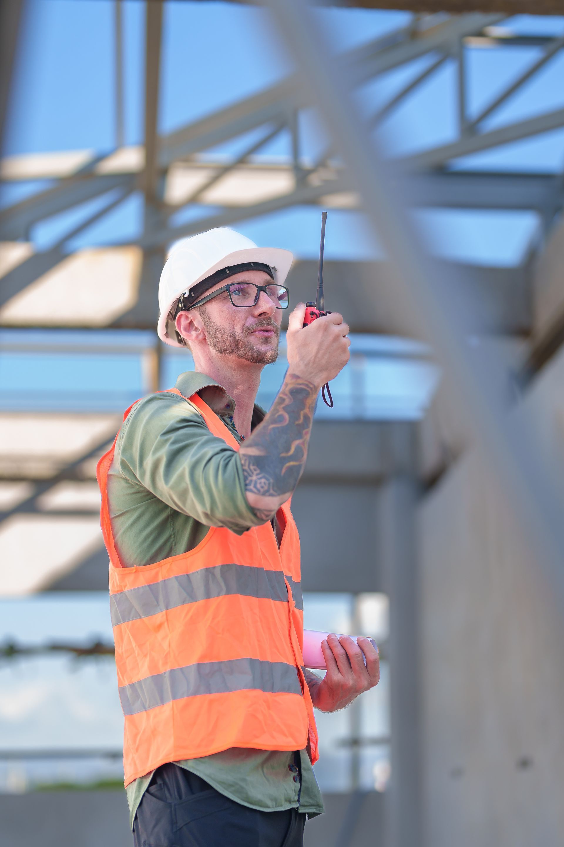 Un ouvrier du bâtiment, portant un casque blanc et un gilet orange, utilise un talkie-walkie sur un chantier.