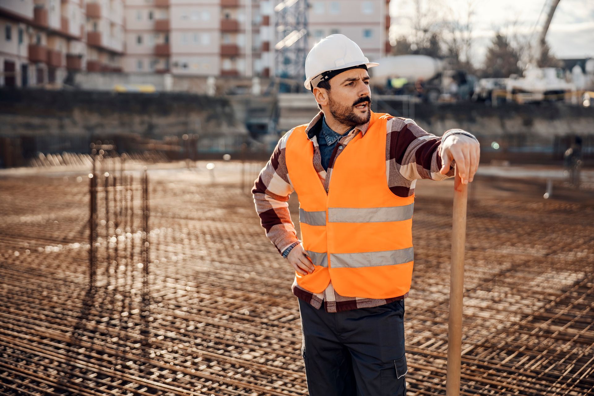 Un ouvrier du bâtiment, portant un gilet de sécurité et un casque, s'appuie sur une pelle sur un chantier.