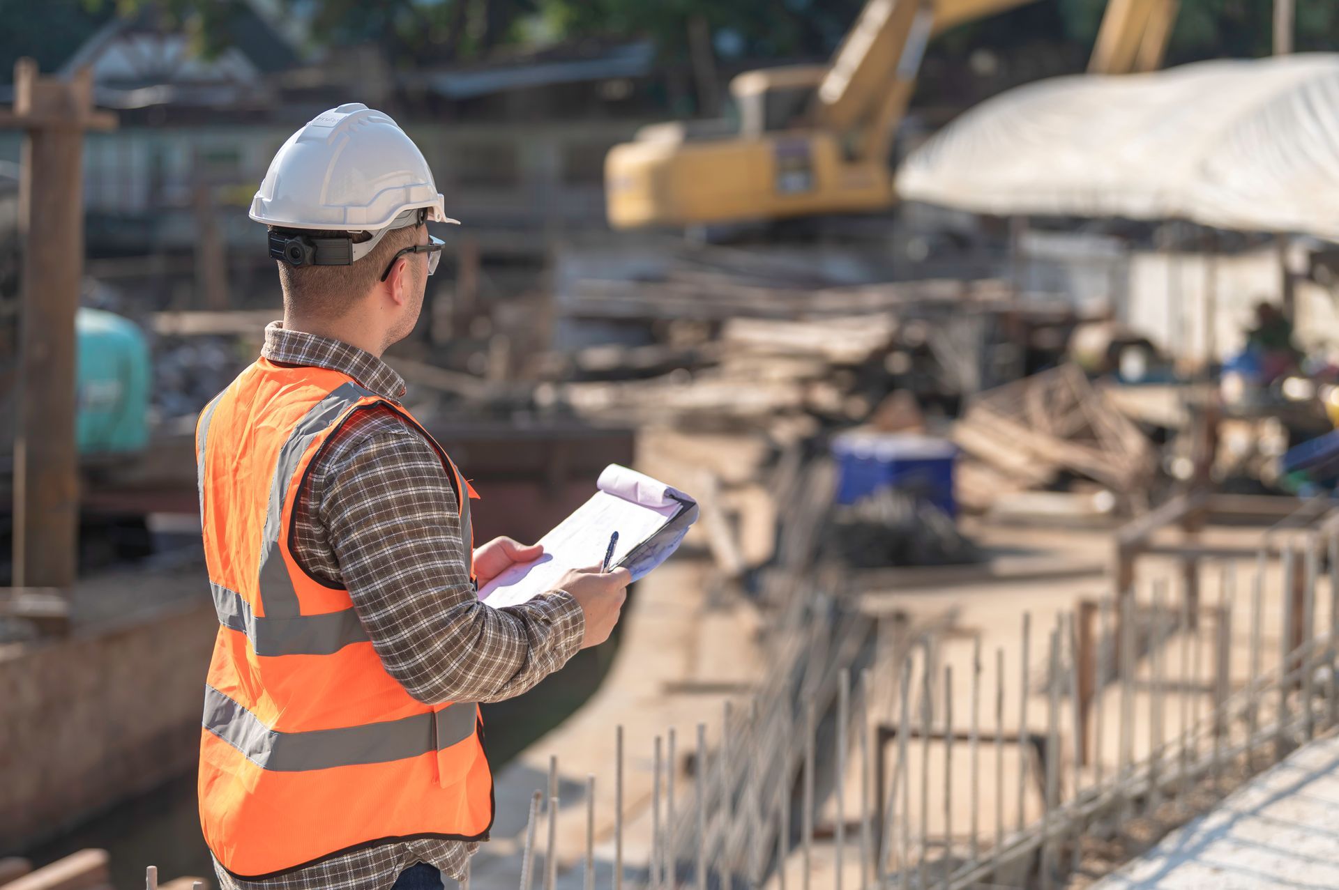 Un ouvrier du bâtiment en gilet orange et casque de chantier consulte un tableau sur un chantier.