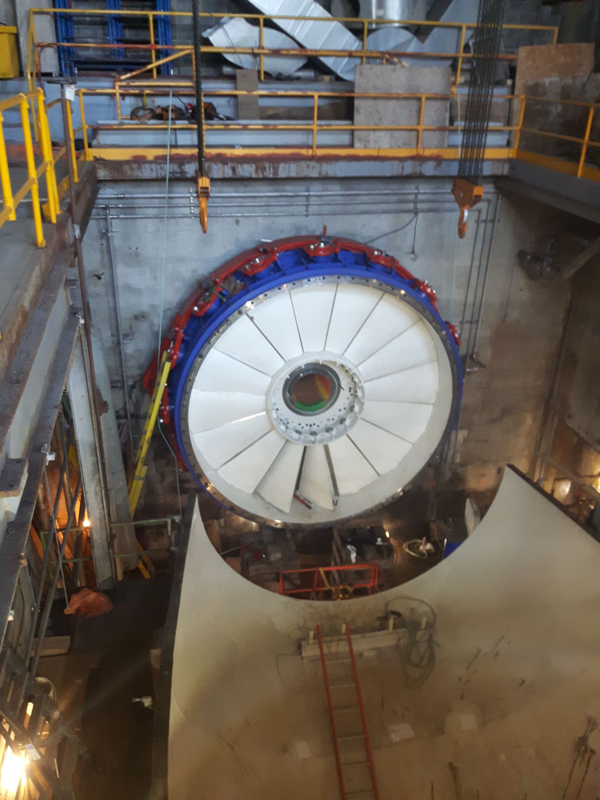 Grande turbine à pales blanches, anneau bleu et rouge, à l'intérieur d'une chambre industrielle en béton.