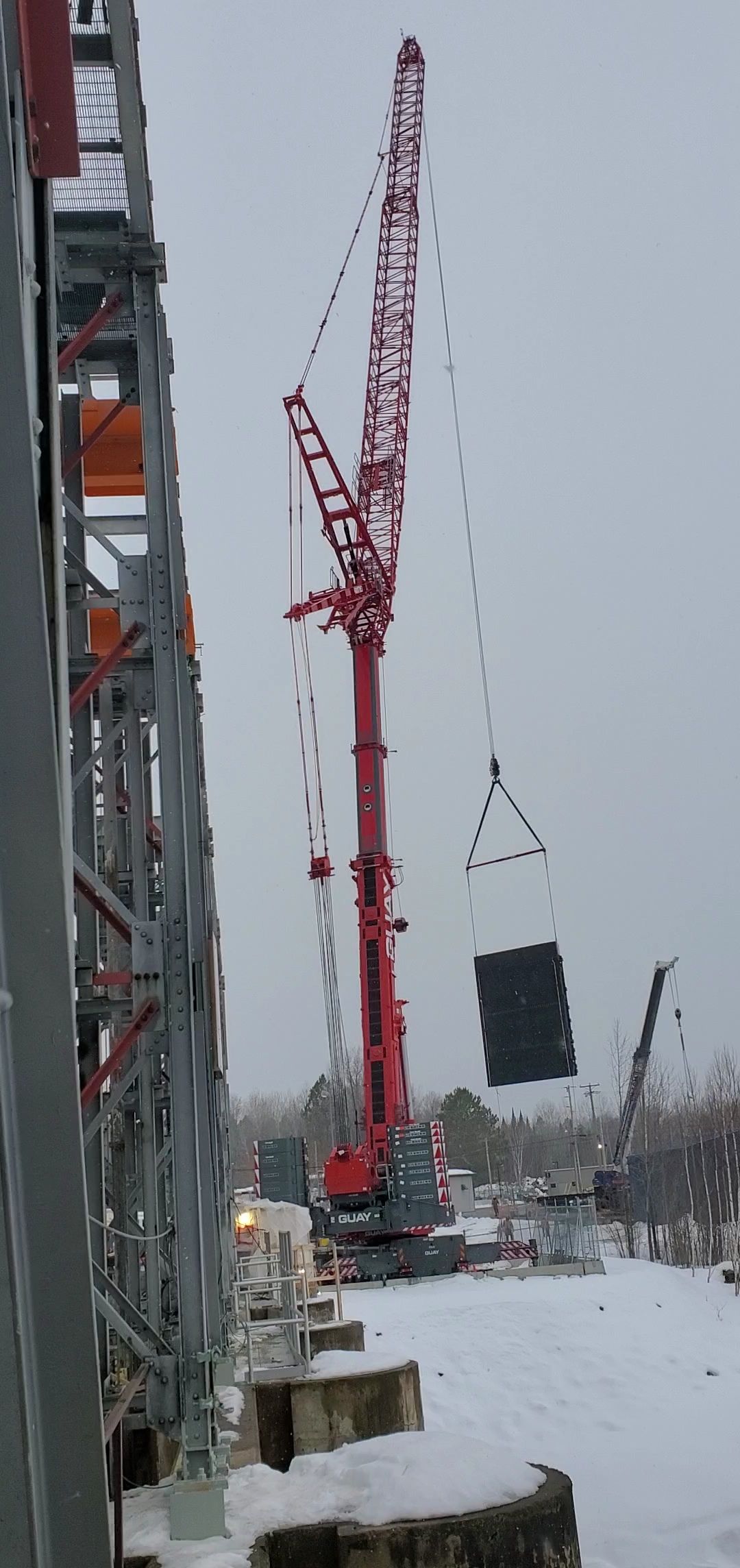 Une grande grue rouge soulève un panneau carré sombre sur un chantier enneigé. Ciel gris.
