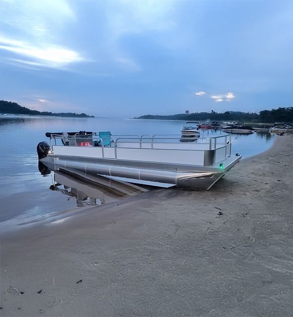 Un bateau ponton en aluminium est échoué sur une plage de sable au crépuscule. Le ciel est couvert et l'eau est calme.