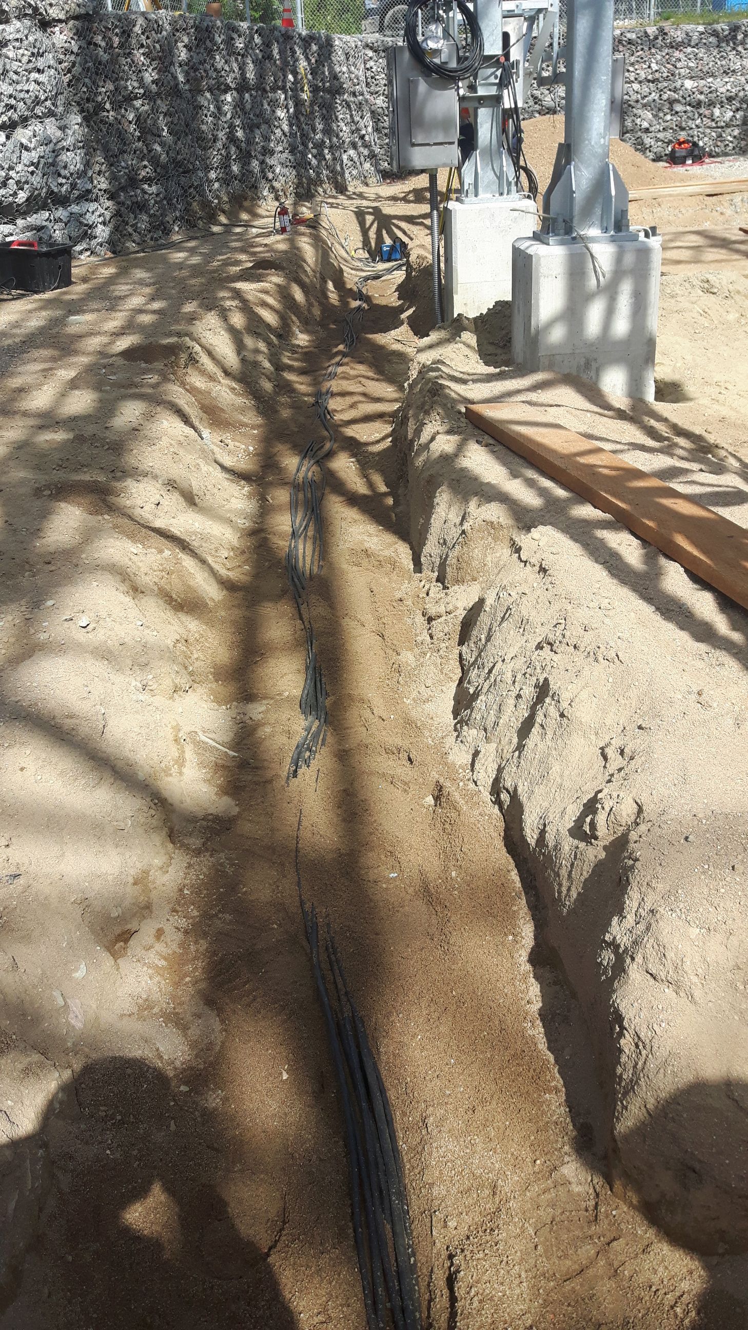 Tranchée creusée dans la terre, tuyau noir visible, près d'une base en béton et d'un mur en pierre.