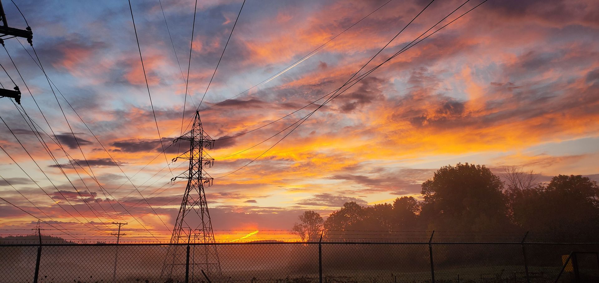 Coucher de soleil sur un champ où des lignes électriques et une clôture en grillage se découpent sur le ciel coloré.