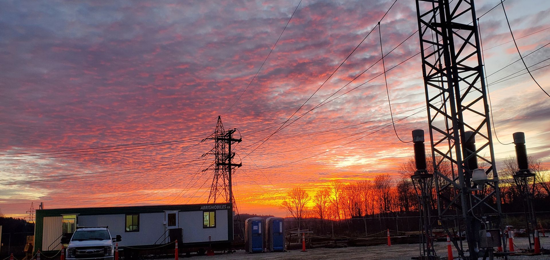 Coucher de soleil spectaculaire sur des poteaux électriques, des lignes électriques, un bâtiment et des toilettes portables ; le ciel s'embrase de teintes orangées et rouges.