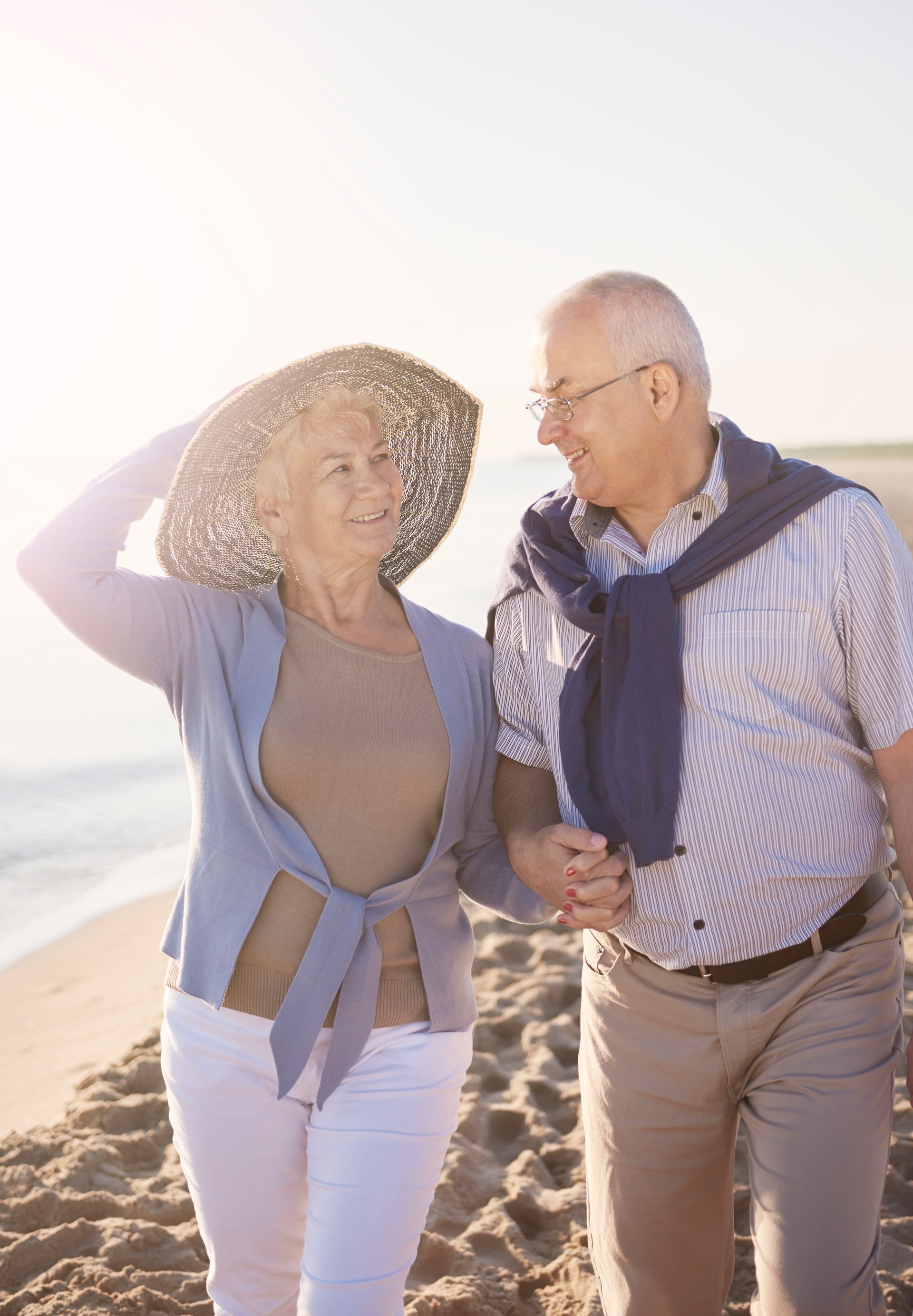 An elderly couple is walking on the beach holding hands.