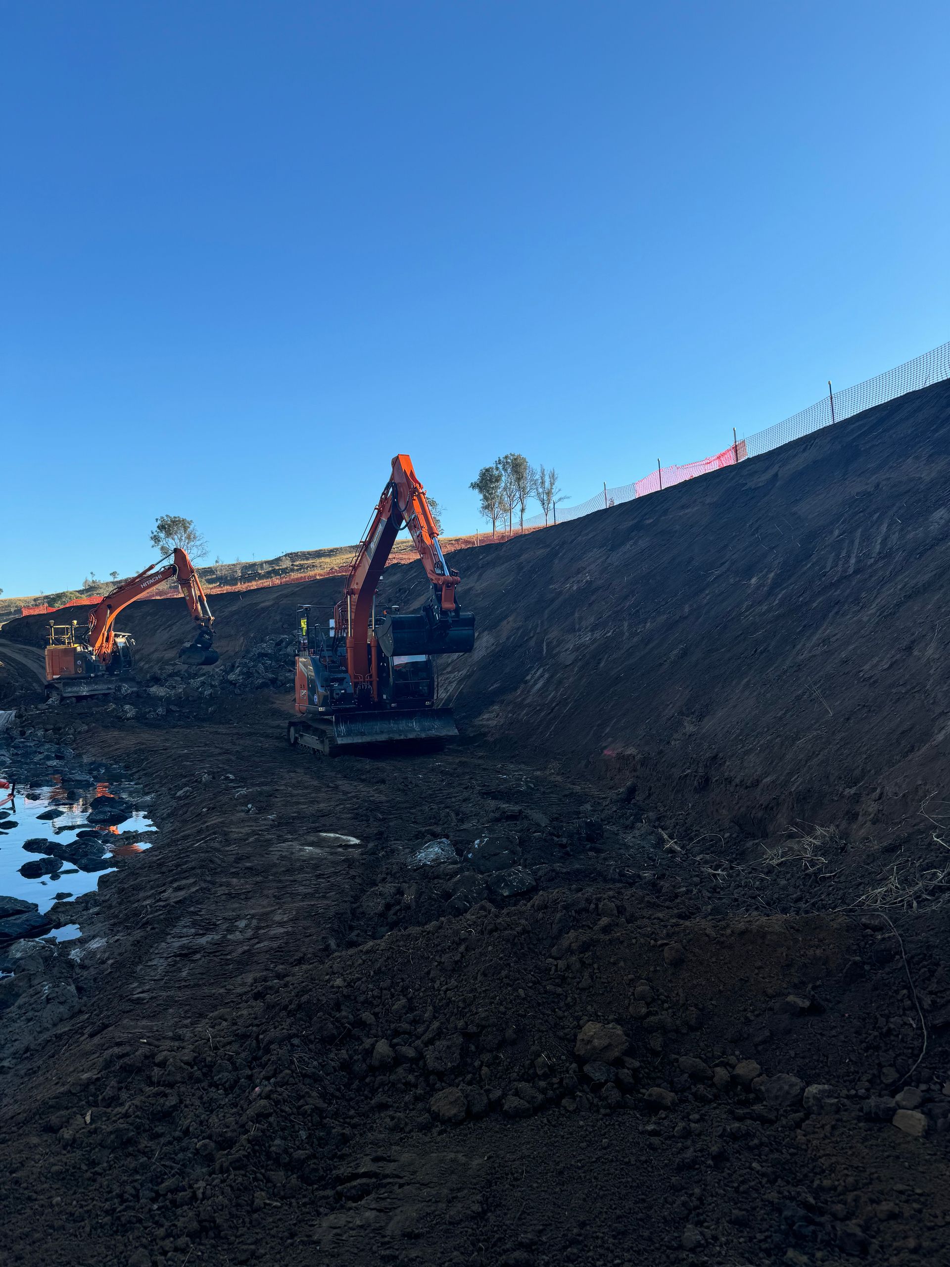 A couple of Spencer Civil & Earthworks excavators are clearing dirt from the base of a hill.