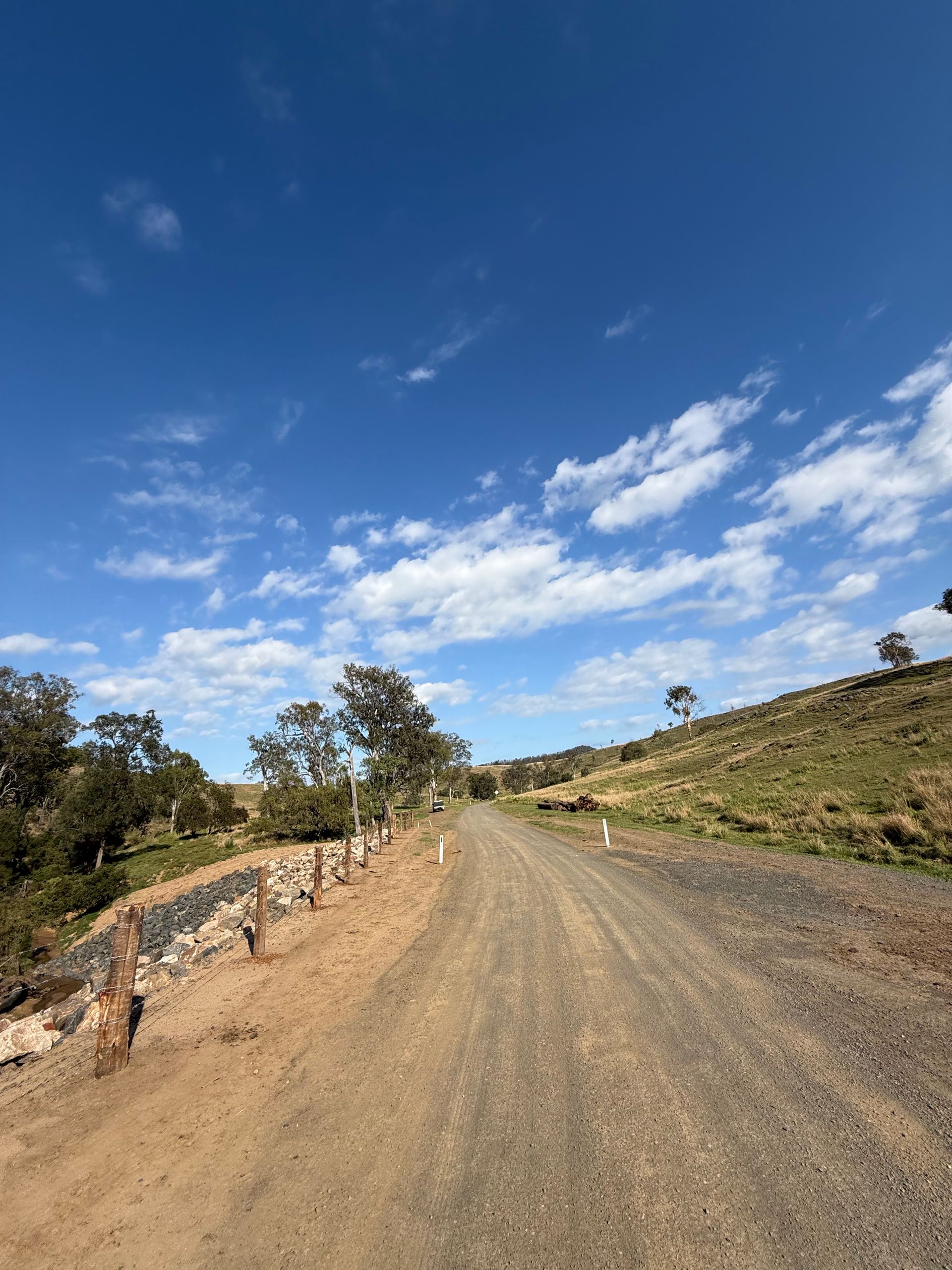 A dirt road with trees on the side and a blue sky, you can see the repaired hillside that was once eroded into a riverbed.