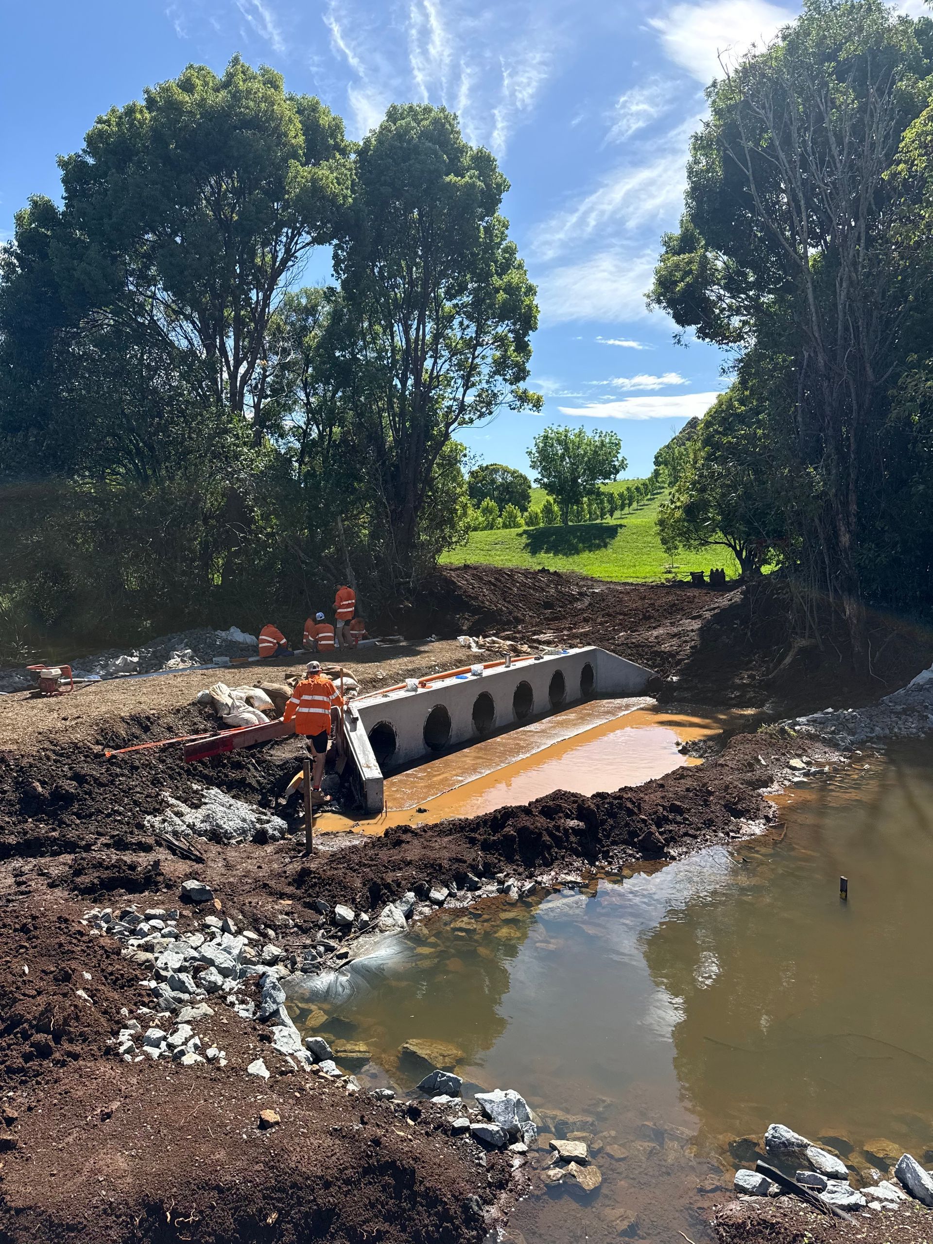 A yellow spencer civil & earthworks excavator is moving dirt next to a silt fence and a body of water.