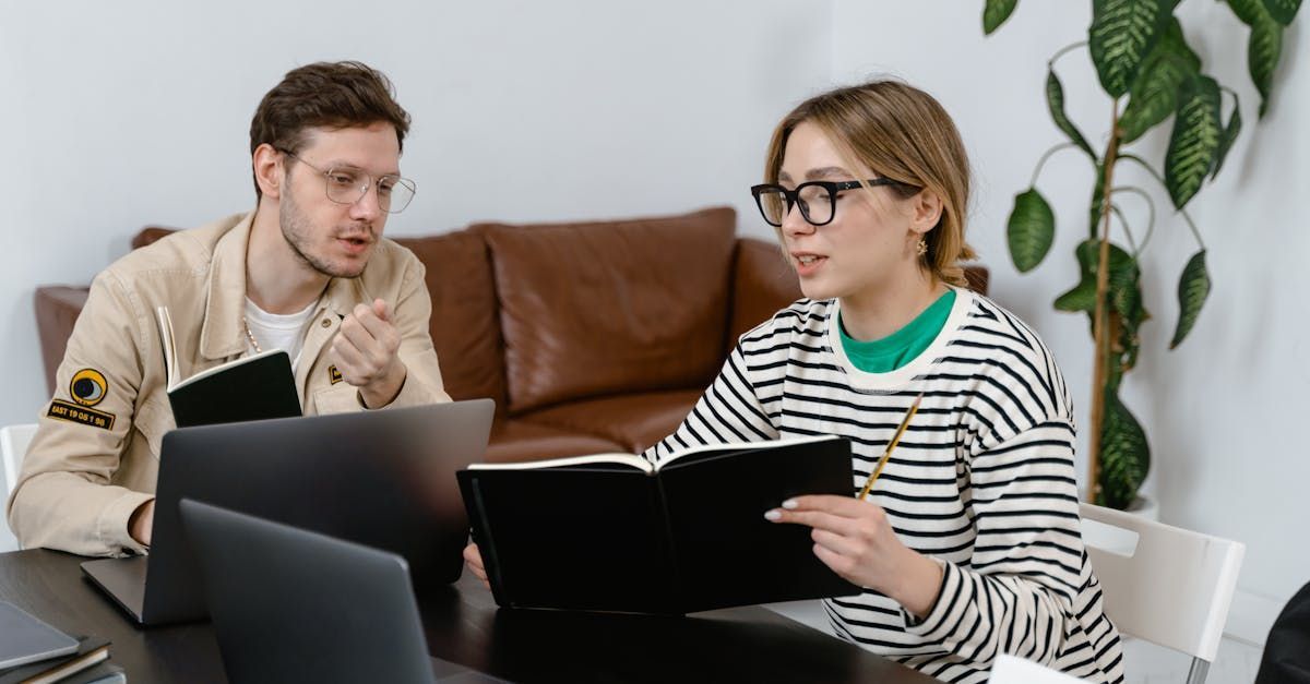 A man and a woman are sitting at a table with laptops and a book.