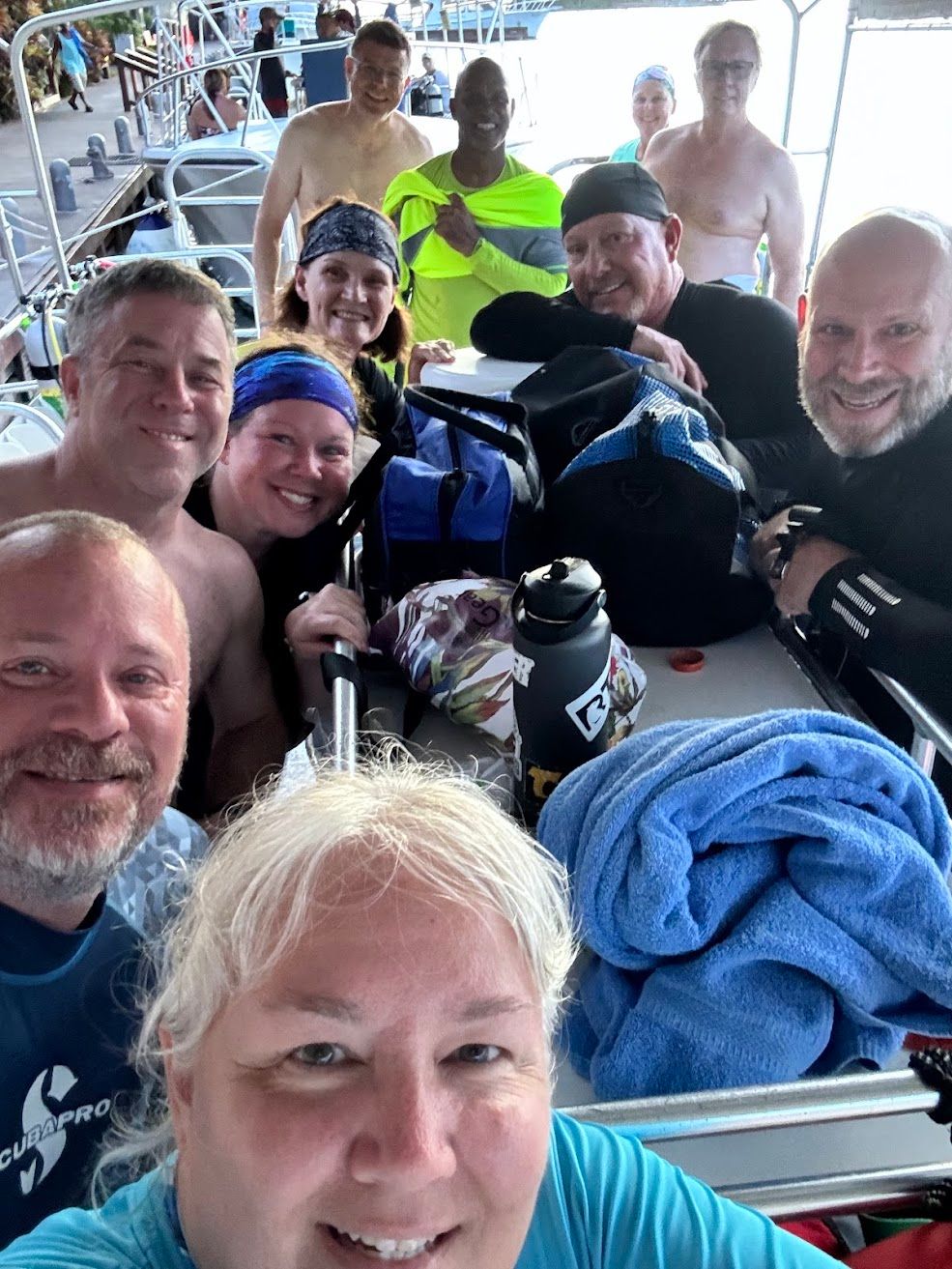 Group of people on a boat smiling. Some are partially clothed, others wearing hats. Blue water and sky in the background.