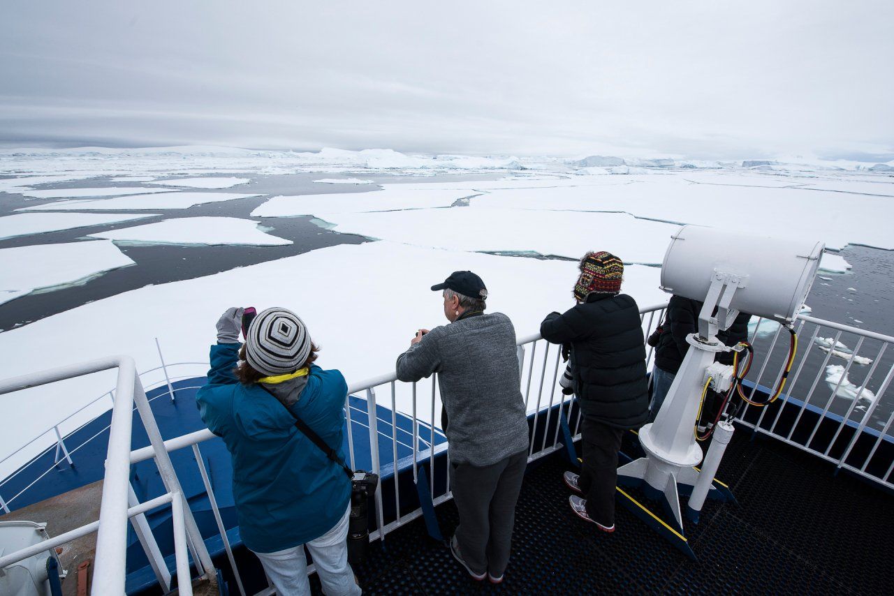 People on ship deck looking at ice floes, one using binoculars. Overcast sky.