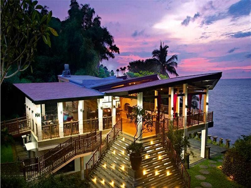Restaurant overlooking the ocean at sunset; wooden deck, illuminated stairs, palm tree silhouette.