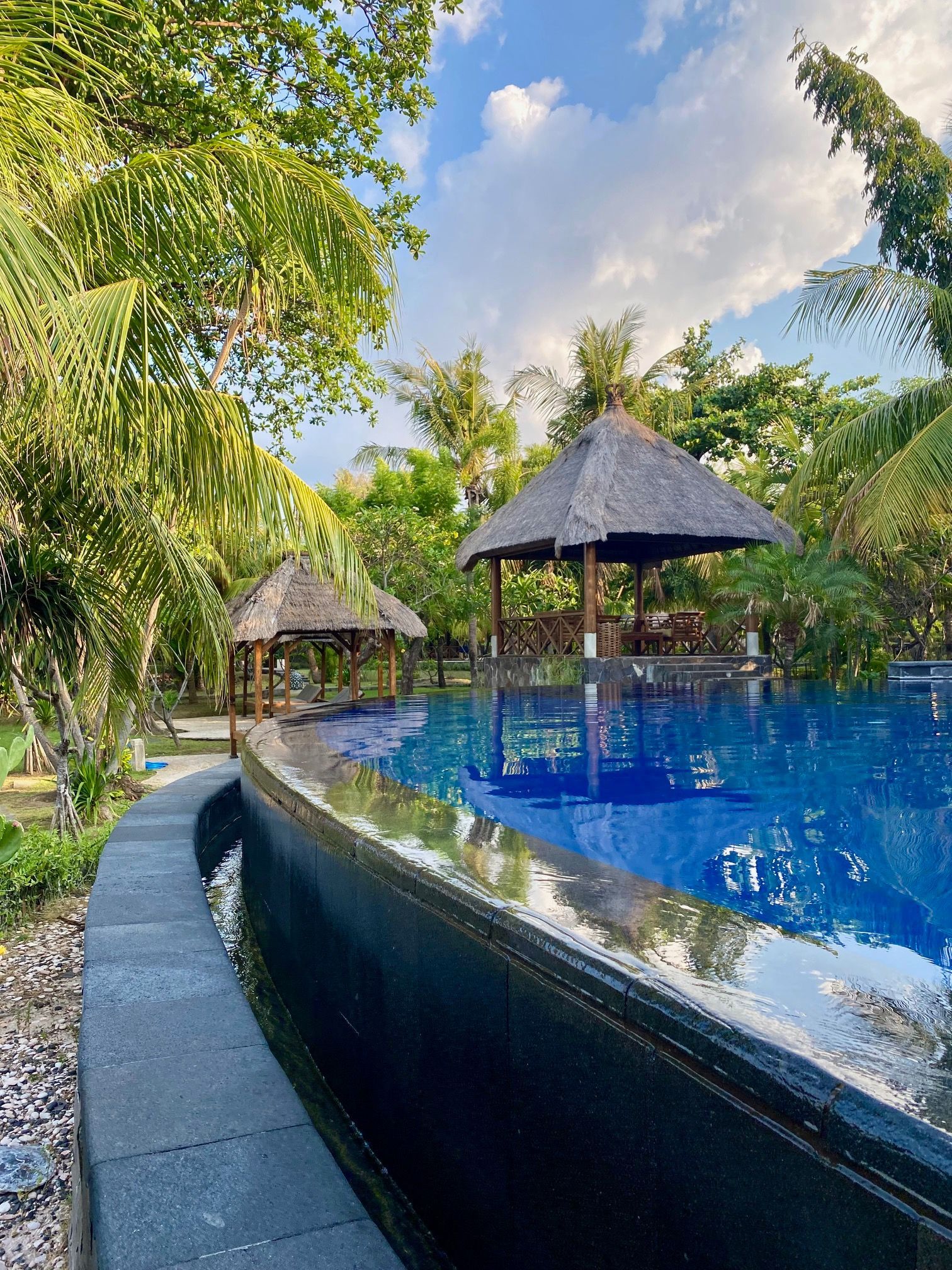 Poolside scene with a blue pool, gray stone border, and two thatched-roof gazebos surrounded by green trees under a partly cloudy sky.