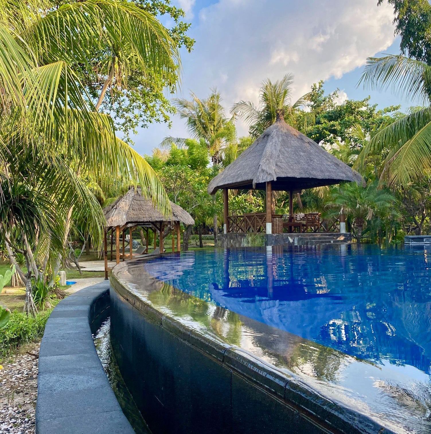 Swimming pool with thatched roof gazebos surrounded by lush greenery under a cloudy sky.