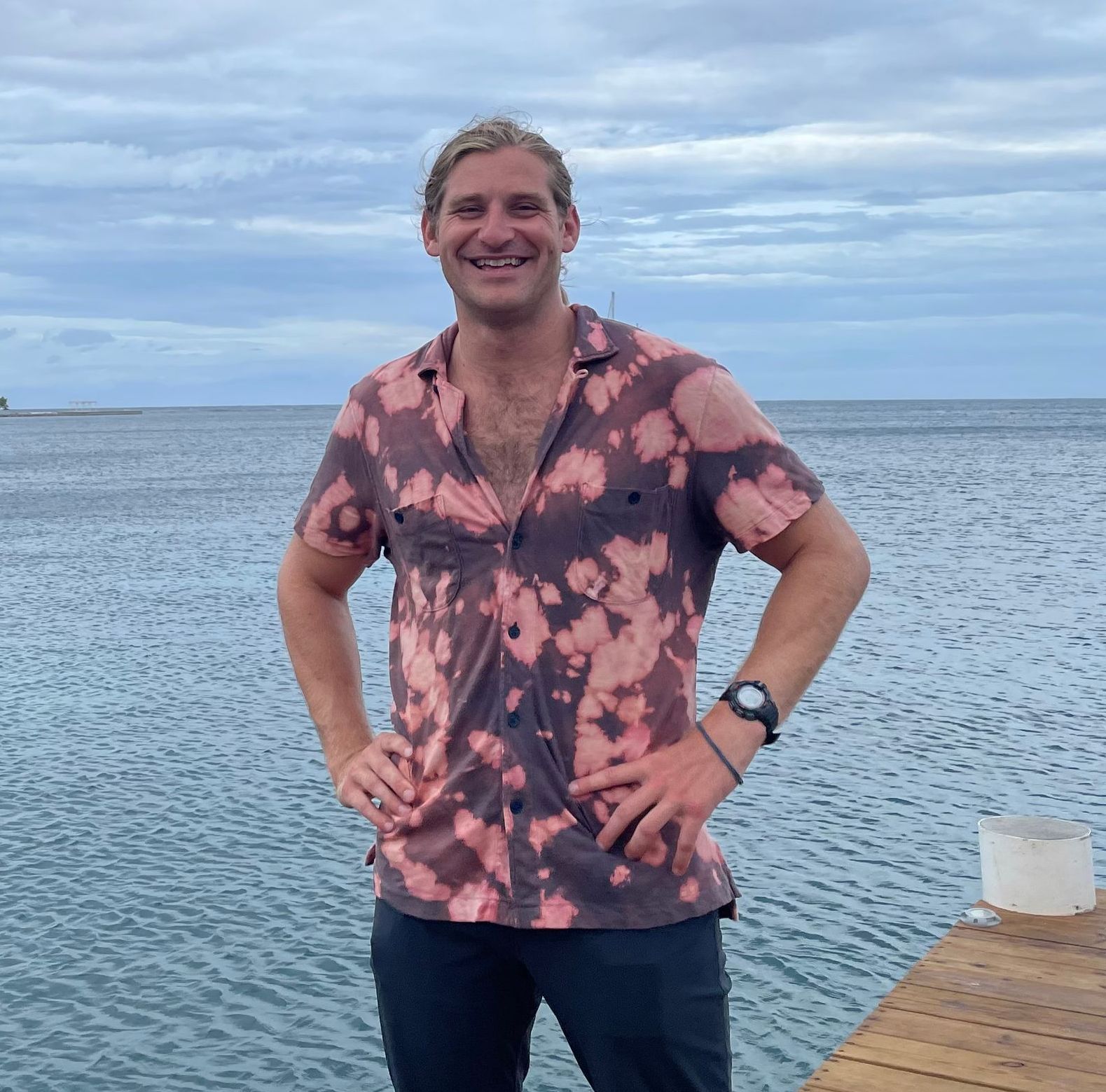 Man in a pink tie-dye shirt stands by water, hands on hips, smiling, wearing a watch.