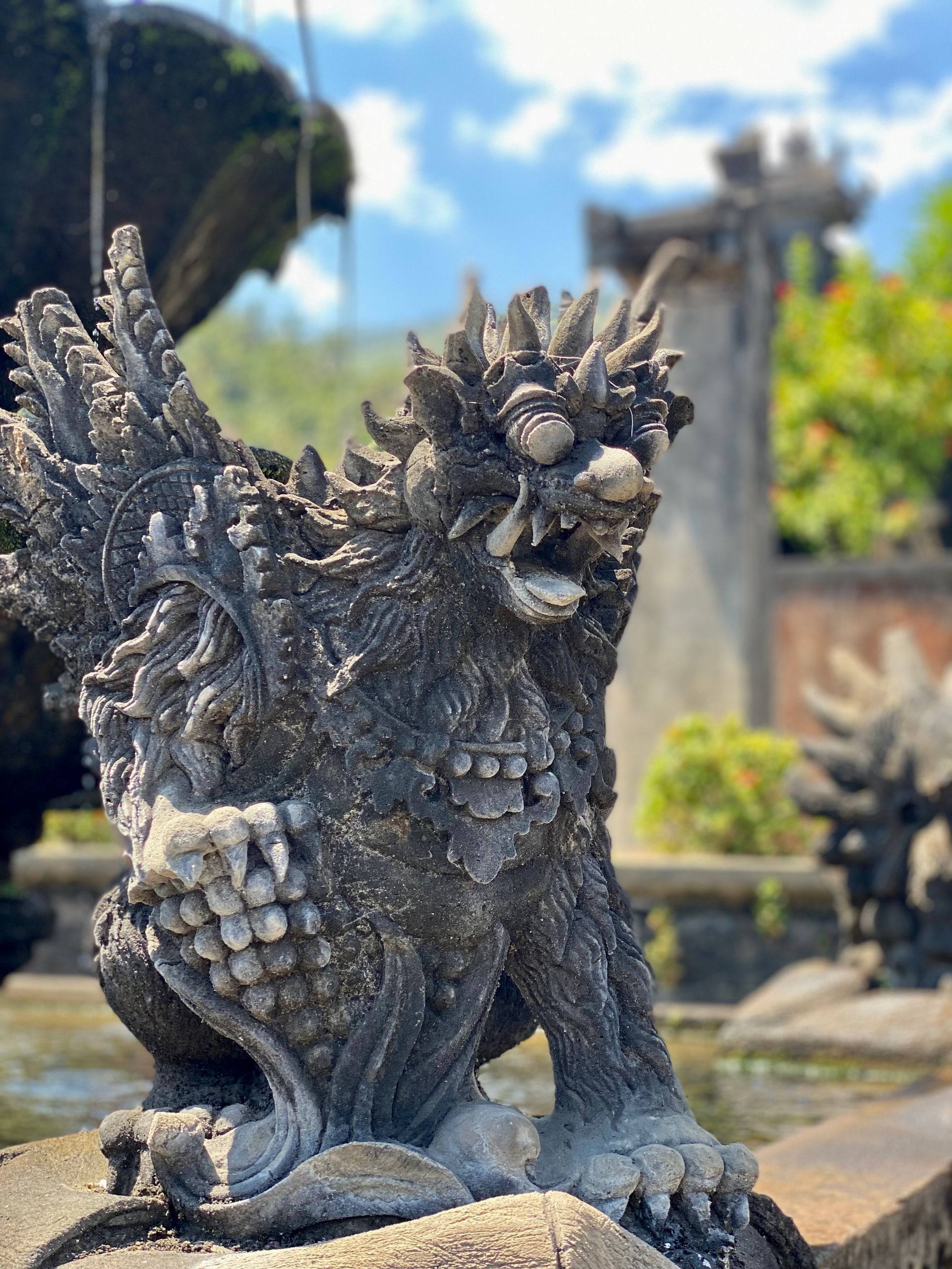 Stone statue of a mythical creature with a winged body and a snarling face, outdoors near water.