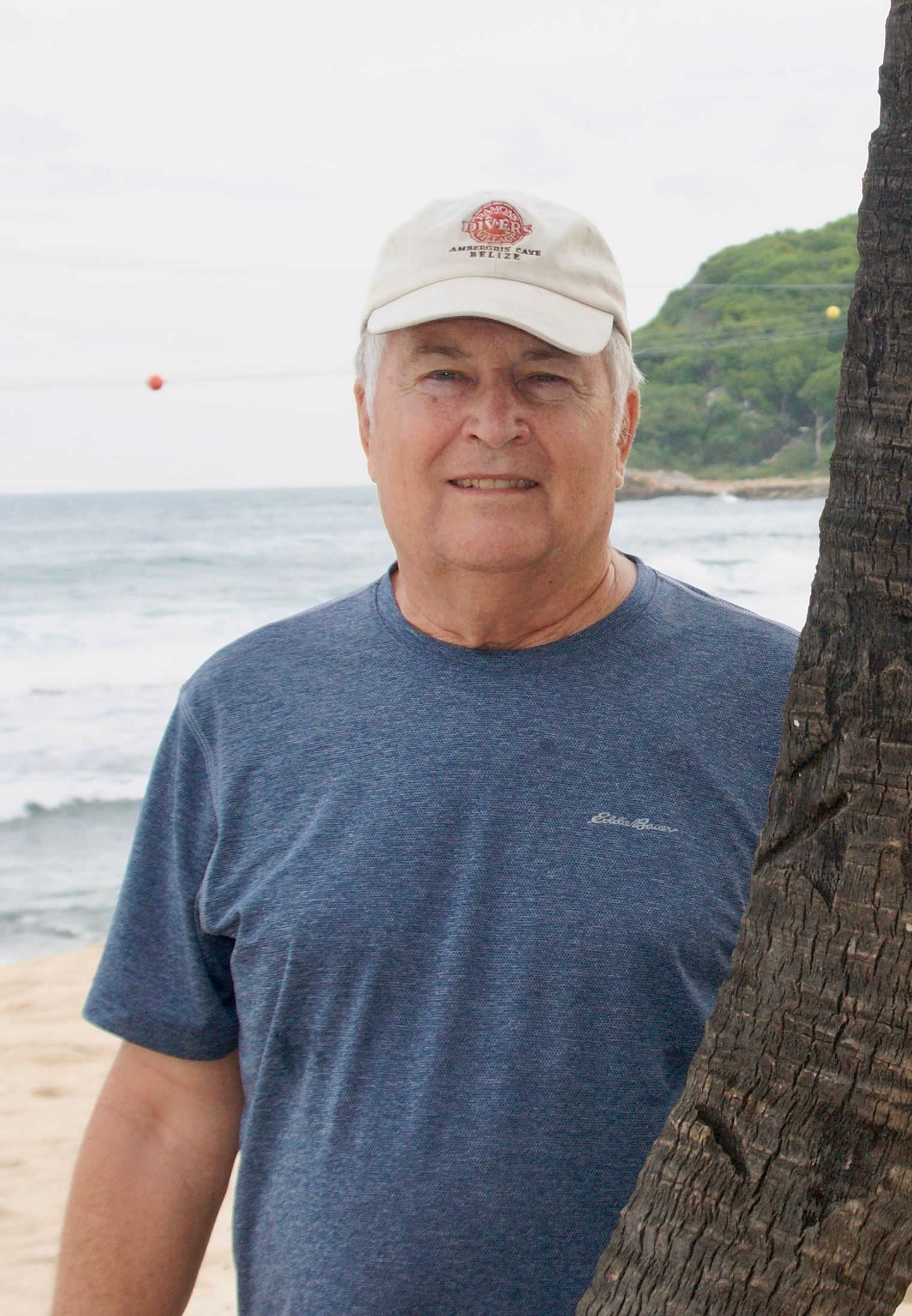 Man in a blue shirt and baseball cap smiles on a beach; ocean and tree trunk visible.
