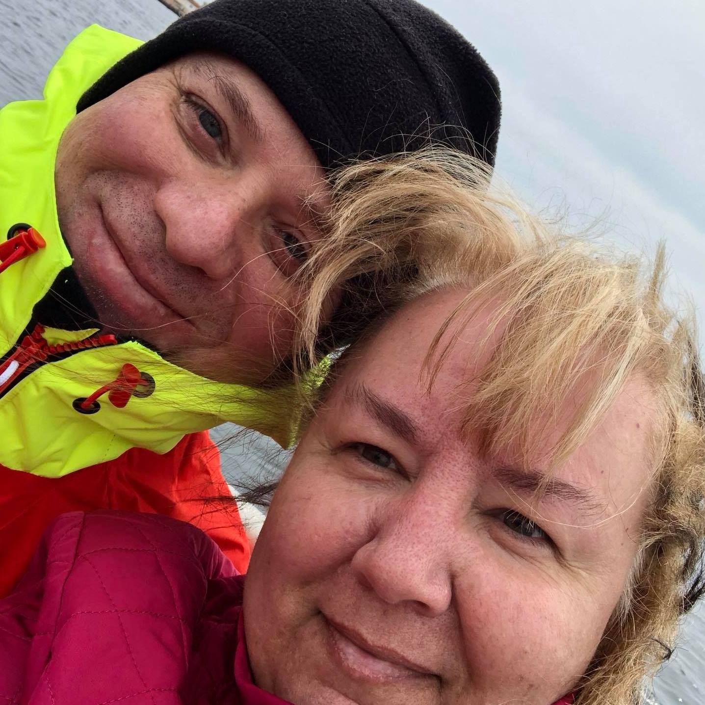 Couple smiling, close-up selfie. Man in black hat and yellow jacket, woman in red, both outdoors.