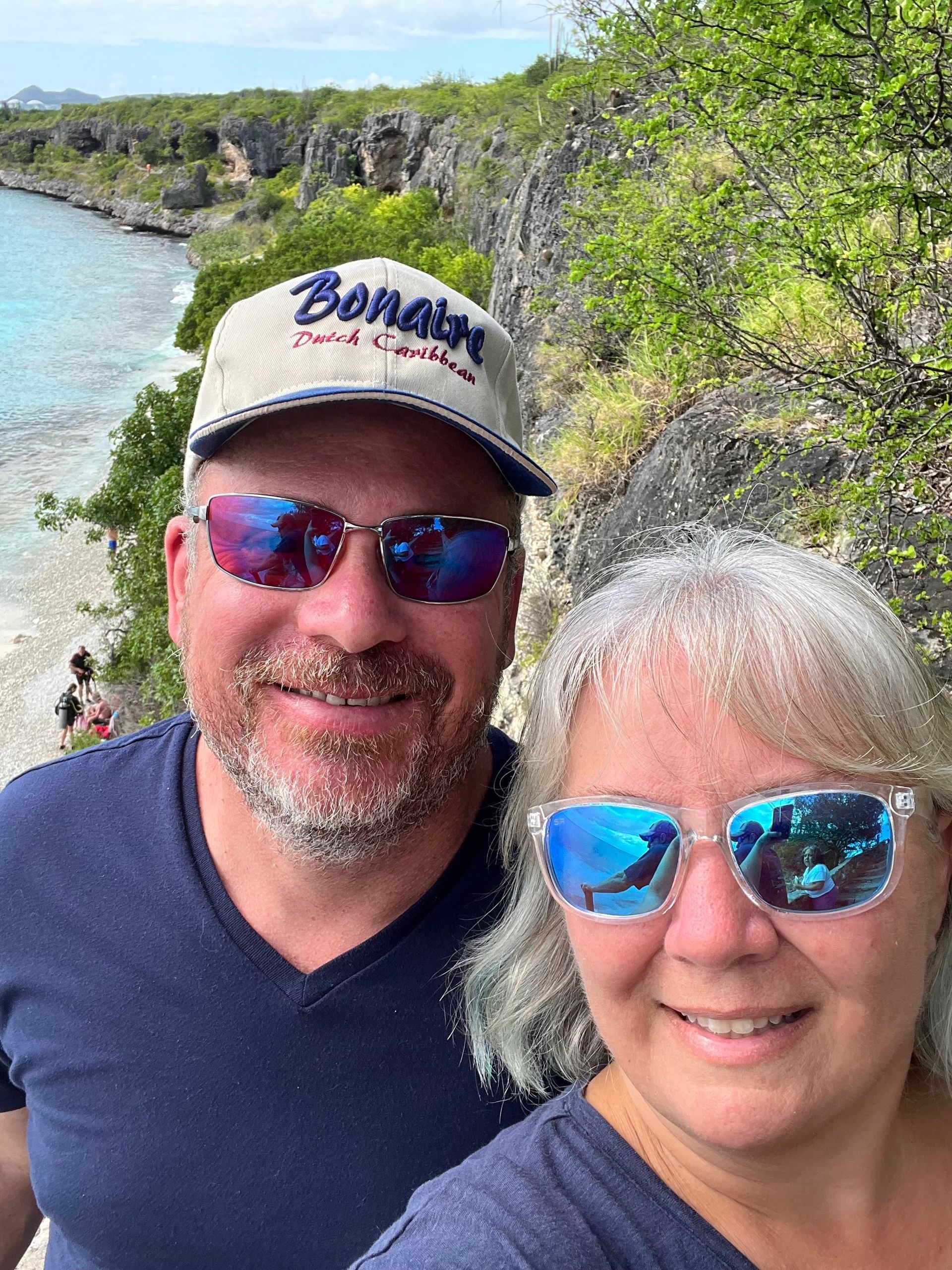 Couple in sunglasses smiling, ocean and cliffs in the background. Man wears a hat.