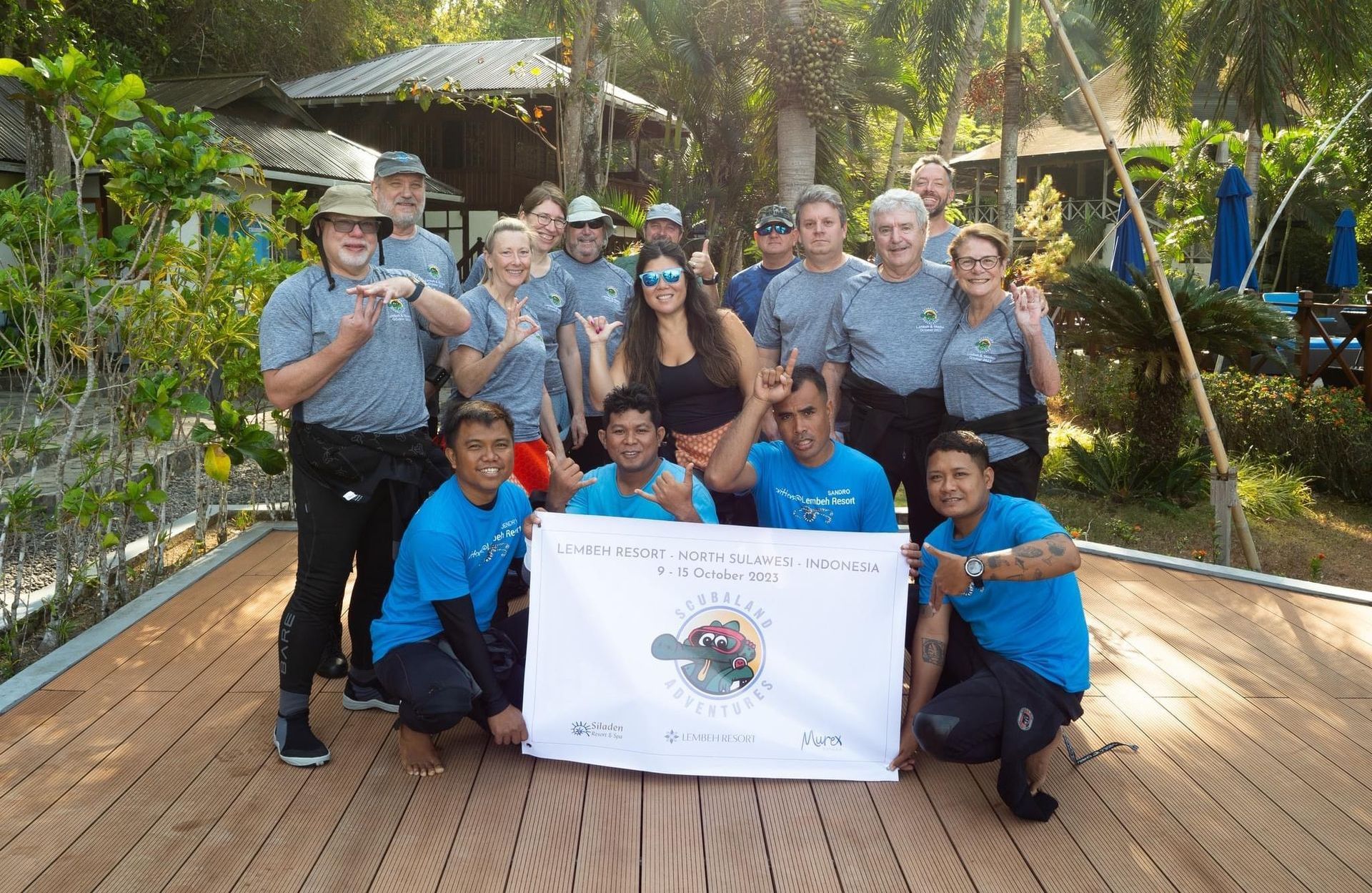 Group of people posing with a banner, some wearing blue shirts and gray tops, outdoors near trees.