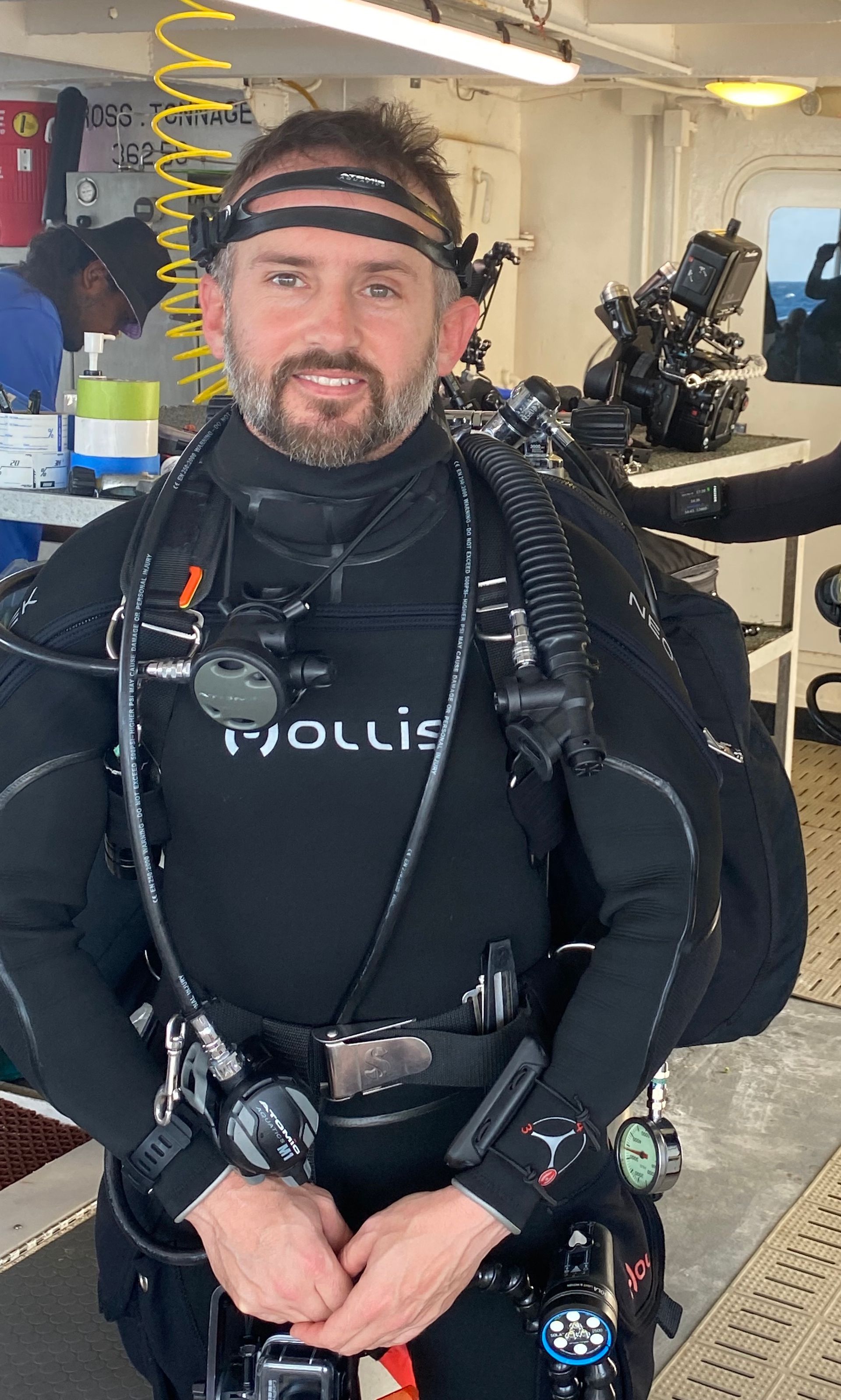 Man in scuba gear, standing inside. Smiling, wearing a wetsuit and equipment, with an underwater environment behind him.
