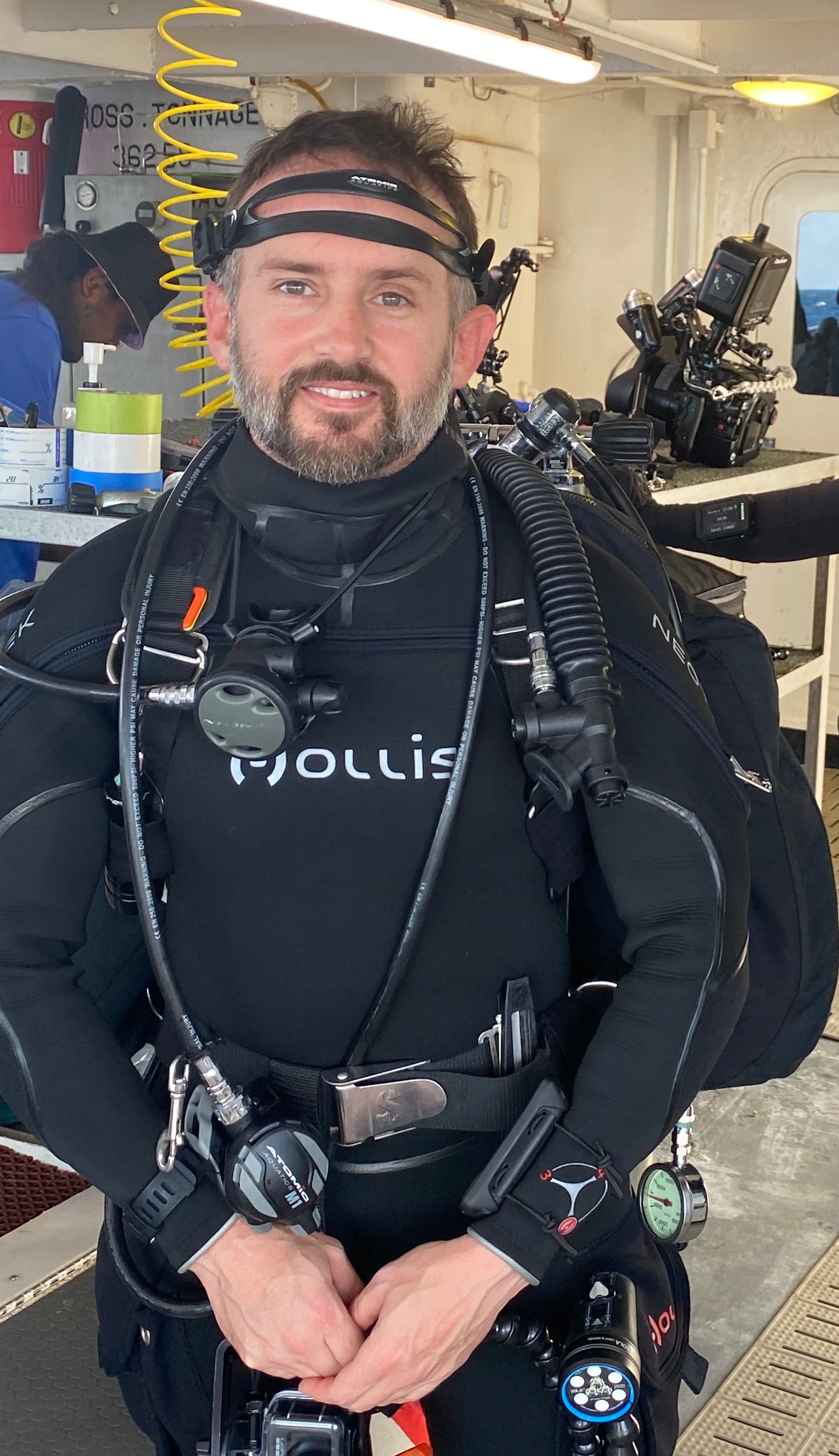 Man in scuba gear, indoors. Wearing a wetsuit, face mask, and regulator. Smiling, arms crossed.
