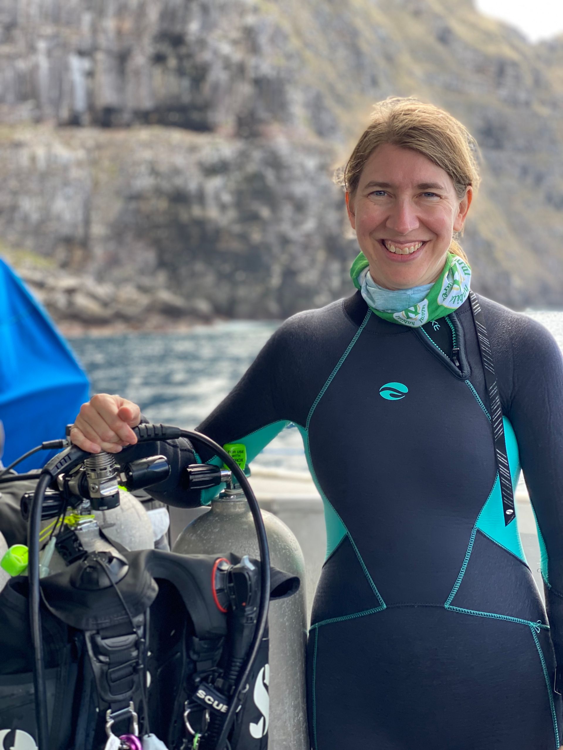 Woman in wetsuit with scuba gear on a boat smiles near rocky shore.