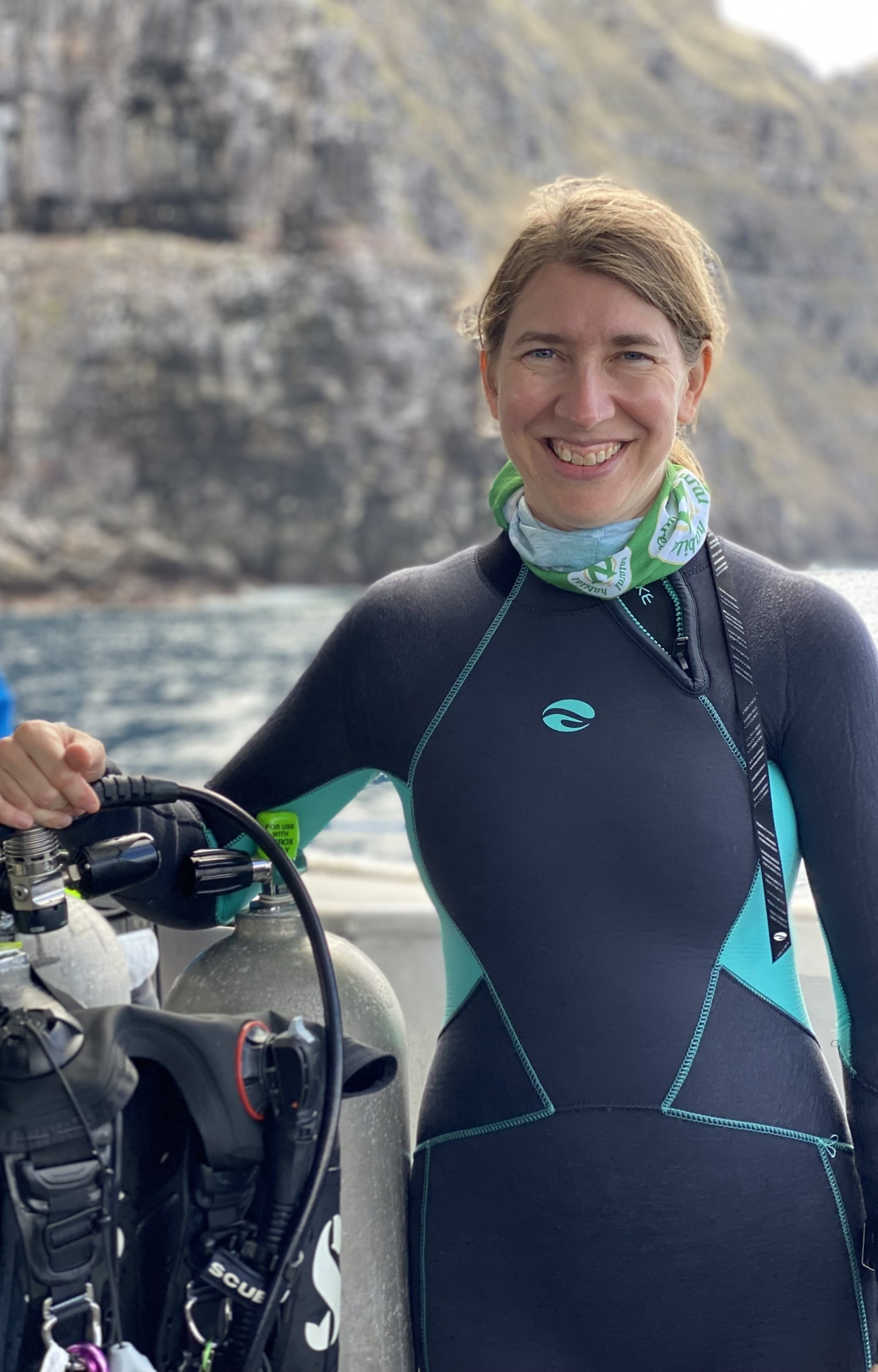 Woman in diving suit smiles, standing near tanks on a boat, with blurred mountainous background.