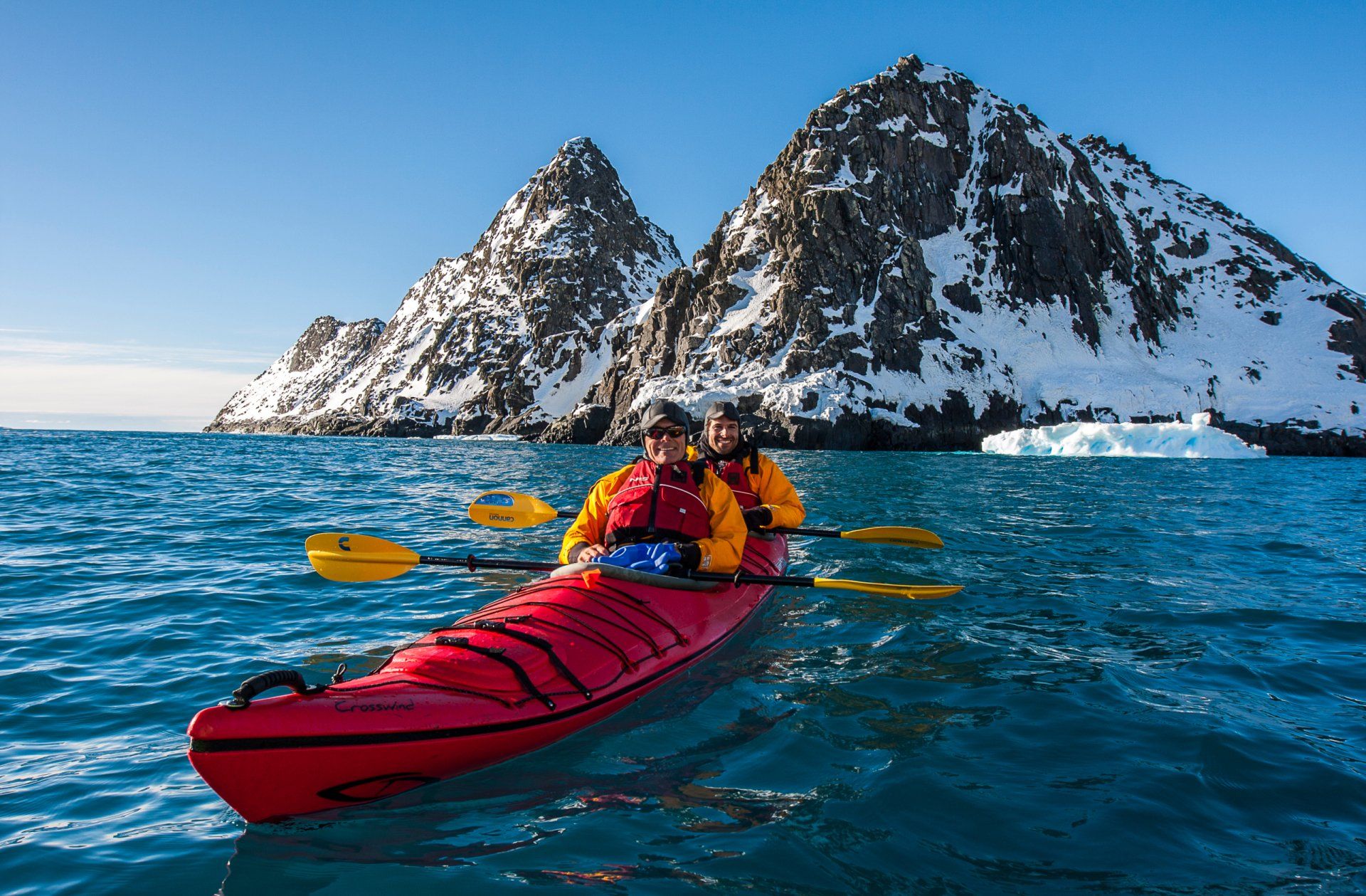 Two people kayaking in a red kayak on blue water, mountains in the background, icebergs, sunny day.