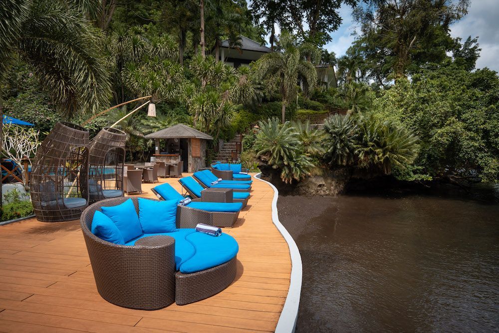 Poolside lounge area with blue cushions, brown wicker furniture, and lush green foliage.