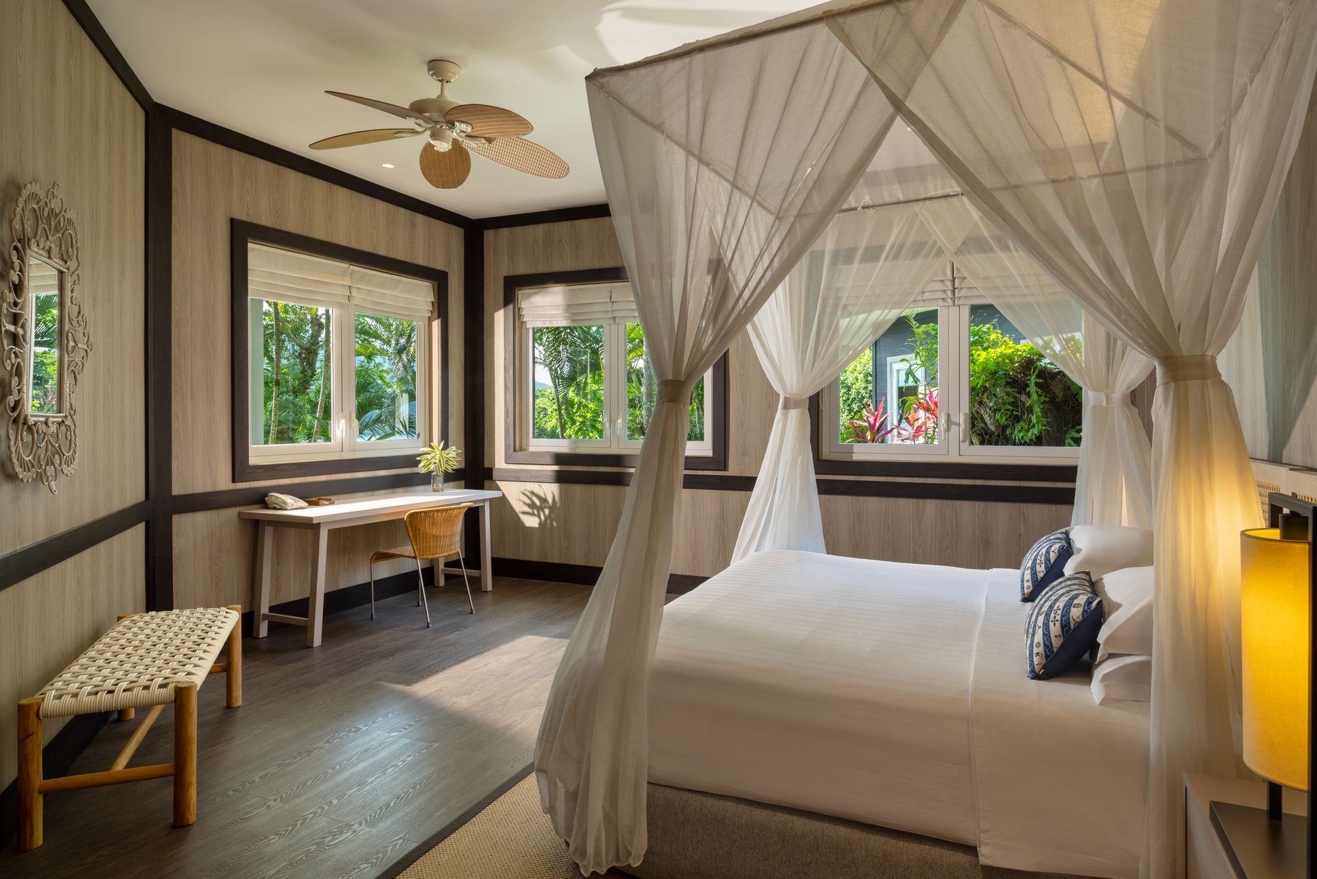 Bedroom with canopy bed, desk by windows, wooden floors and walls. Sunlight streams in.