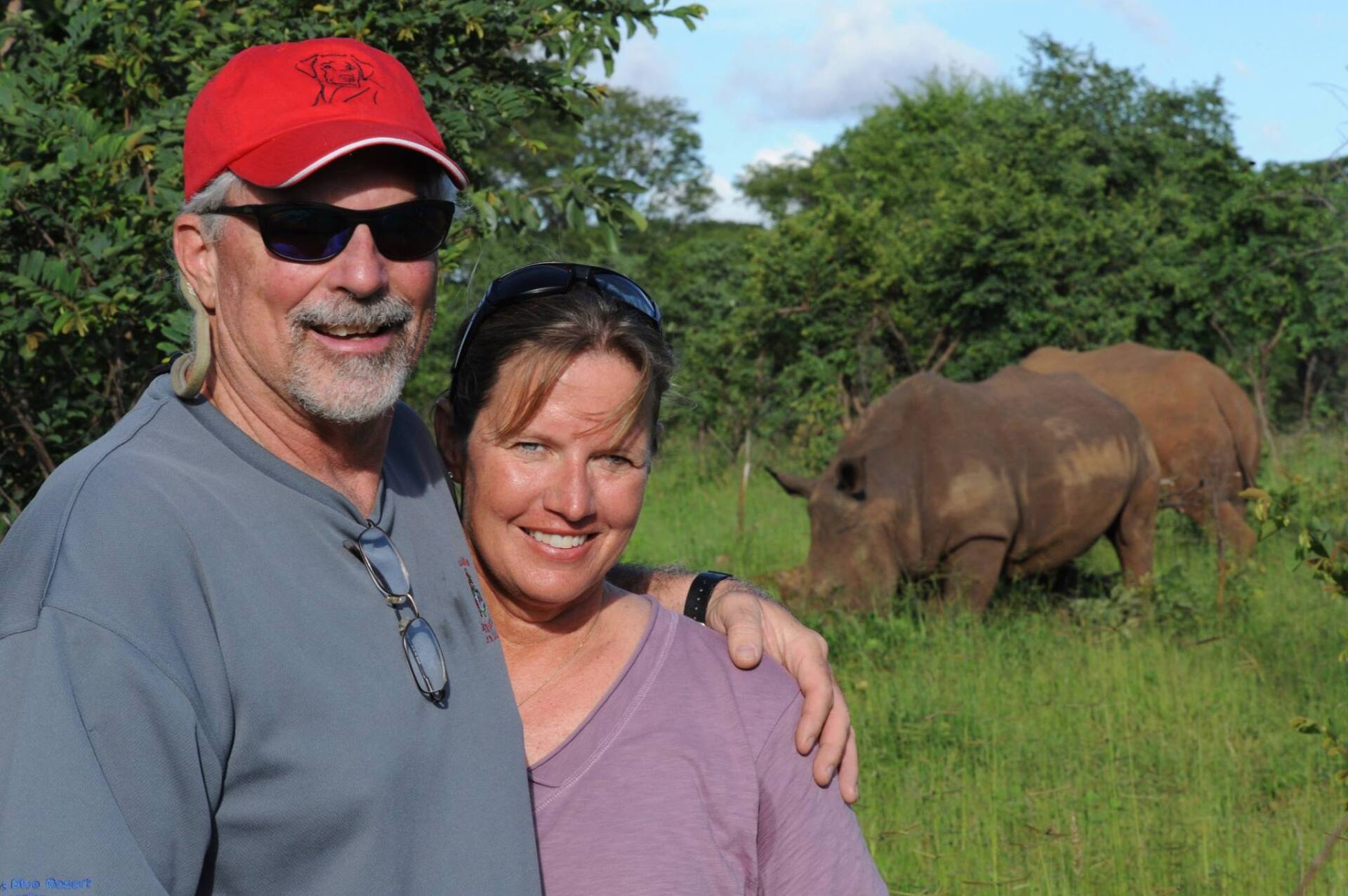 Couple poses with rhinos grazing in a grassy field. The man wears sunglasses and a red cap.
