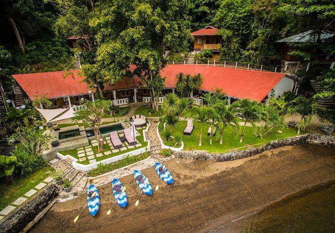 Resort with red roof, small pool, kayaks on shore, surrounded by green trees.