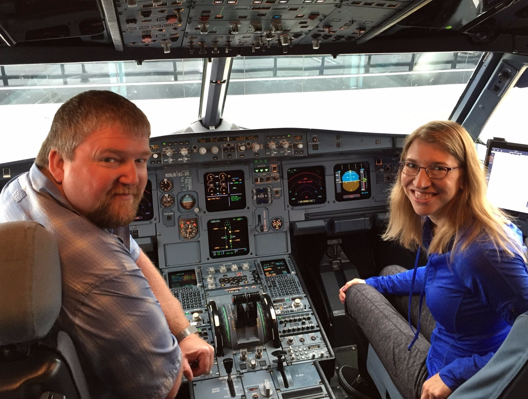 Two people smiling in an airplane cockpit; man on left, woman on right. Bright instrument panel.
