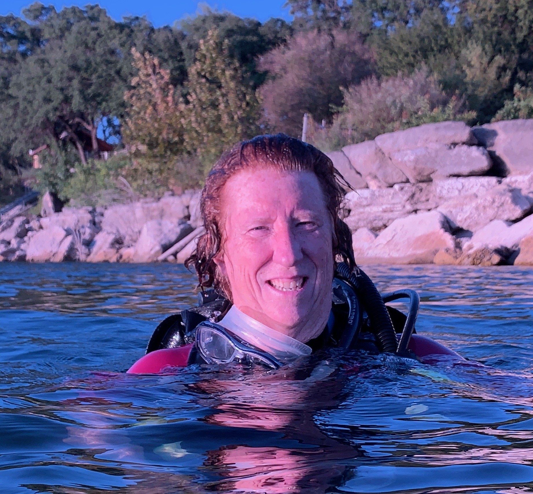 Person in scuba gear in water, smiling, trees and rocks in background.