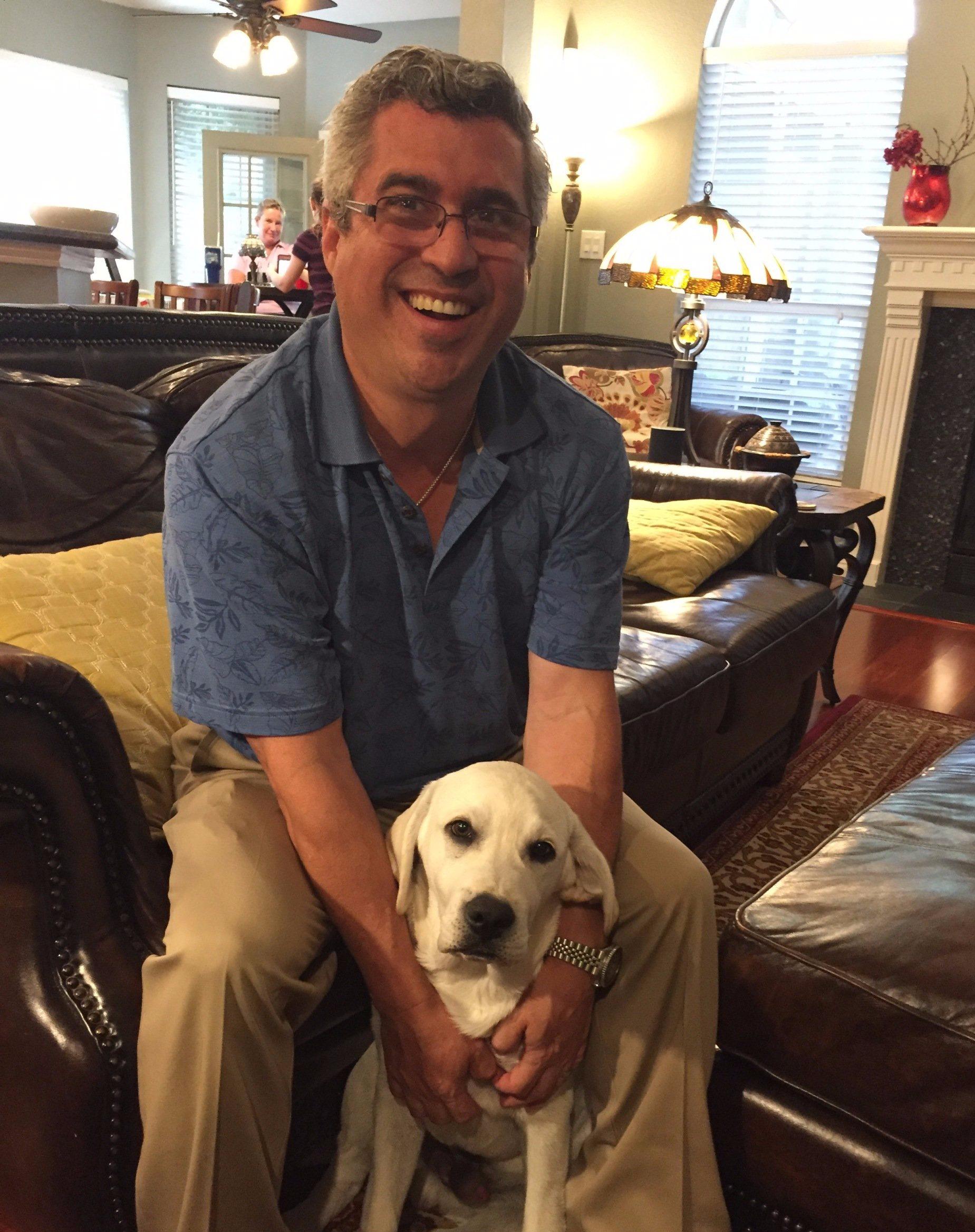 Man sitting on a leather couch, smiling, holding a white Labrador puppy. Home interior with lamp, fireplace.