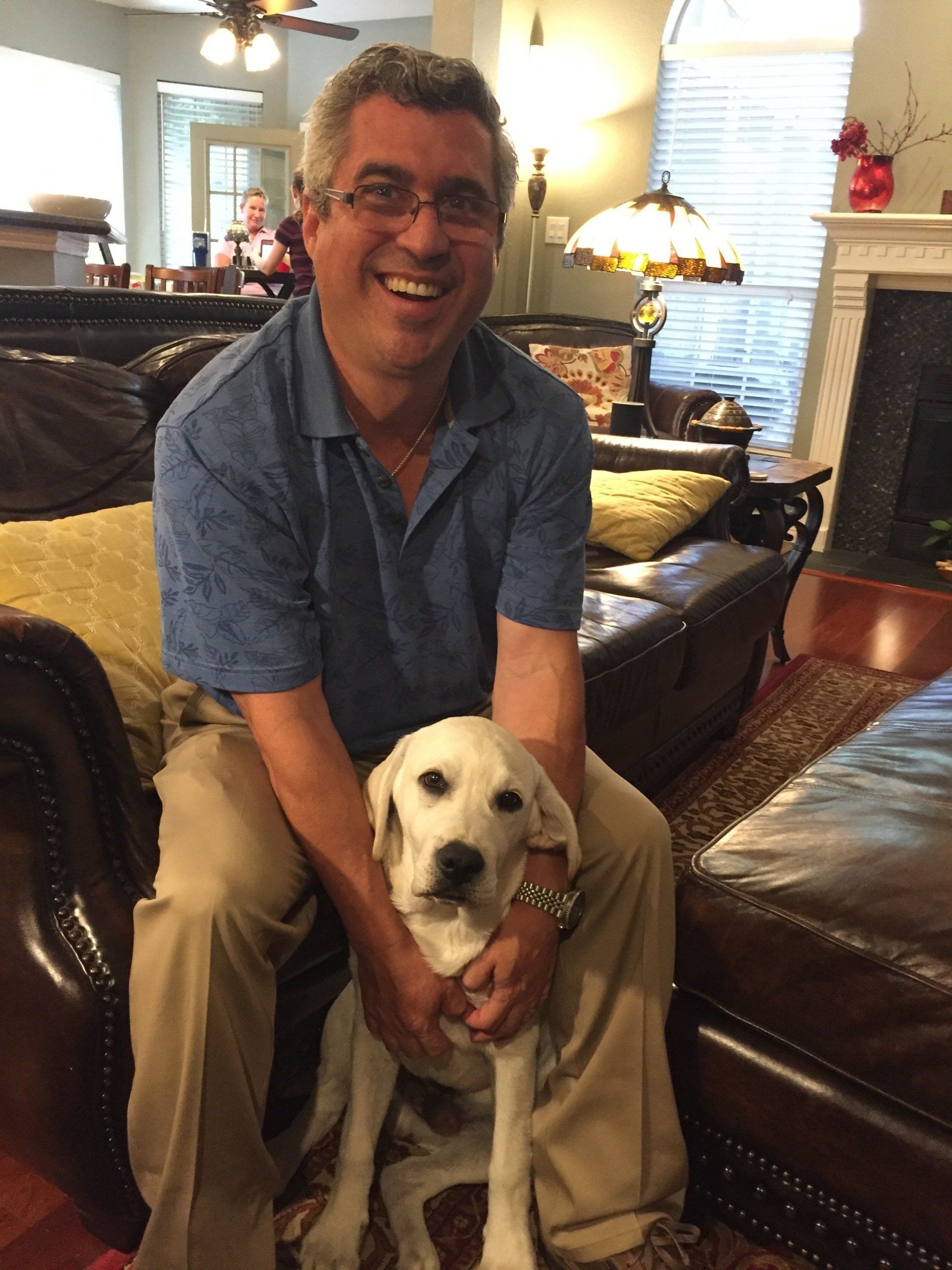 Man sitting on a brown leather couch, smiling and holding a light-colored dog indoors.