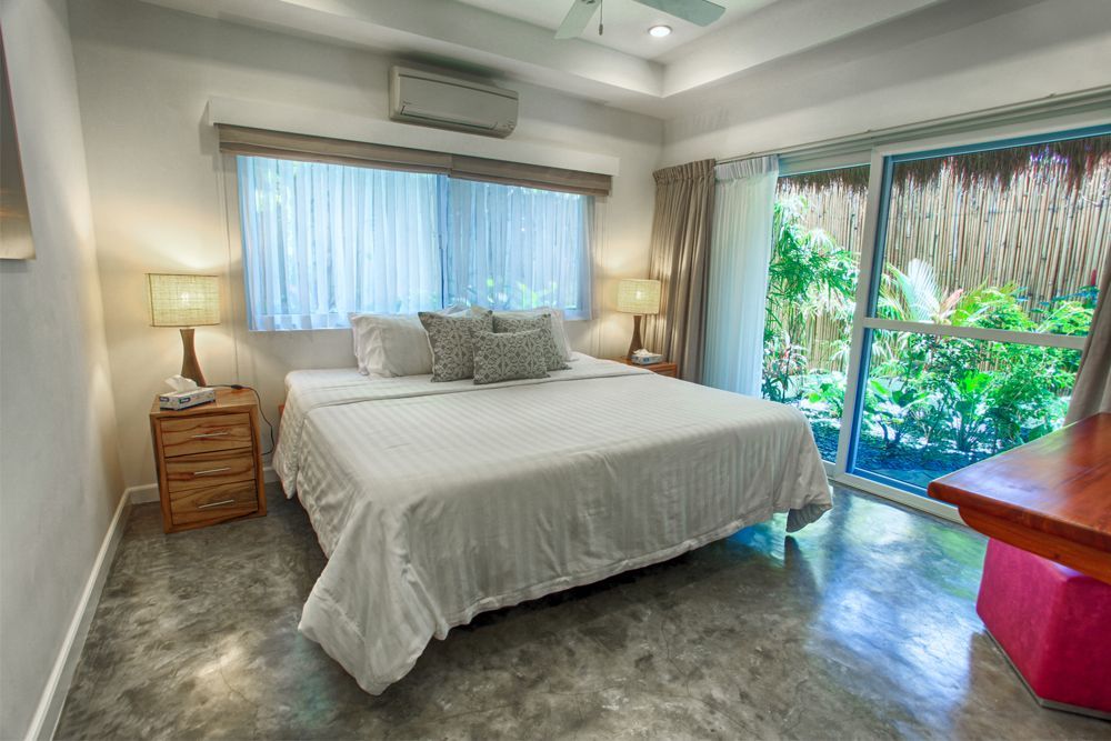 Bedroom with king bed, bedside tables, sliding door to lush green view. White and natural tones.
