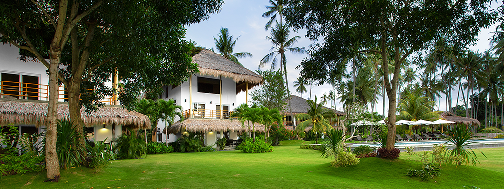 Lush green lawn with white buildings, thatched roofs, and palm trees. Sunny day.